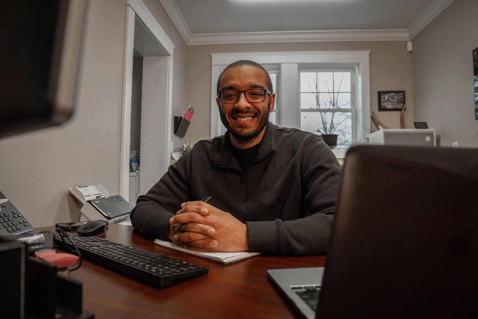 A man with glasses smiling and sitting at a desk with a computer and office supplies in a well-lit room with a window and houseplants in the background.
