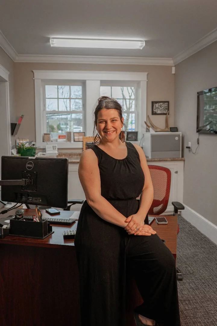 A woman in a black dress standing in an office with a desk, computer, and window in the background.
