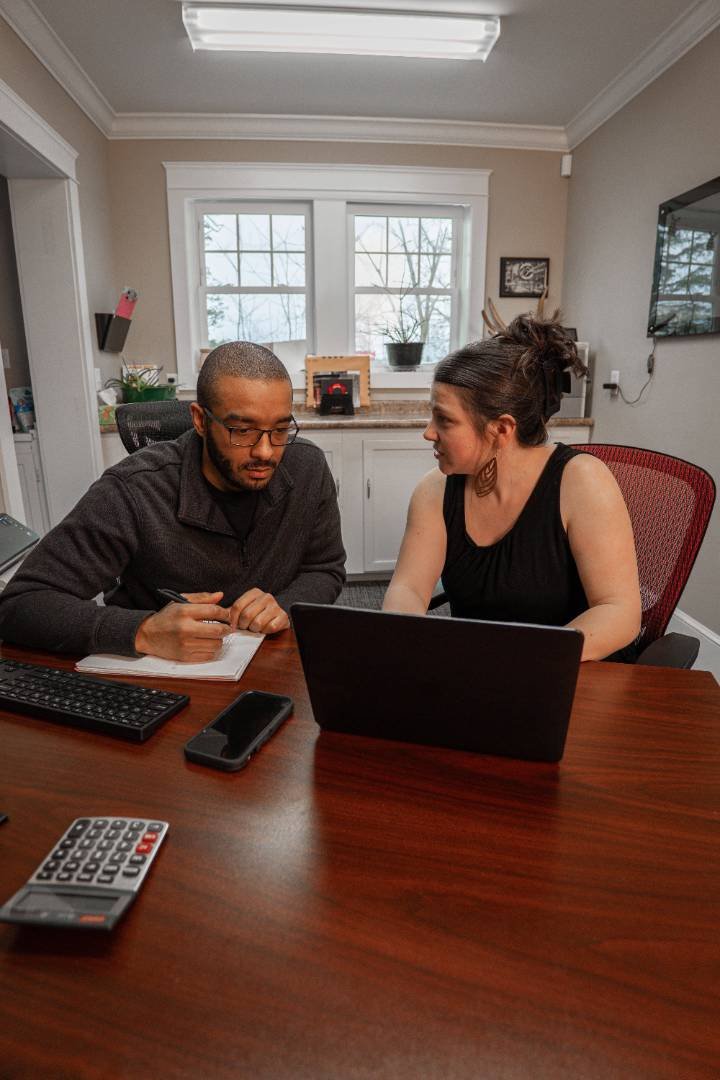 Two people sitting at a wooden table in an office, looking at a laptop. The man on the left is holding a pen and taking notes in a notebook, while the woman on the right is leaning in and engaging with him. There are a keyboard, a calculator, and a smartphone on the table. In the background, there is a window with a view of trees, a desk with some items, and a wall-mounted TV.