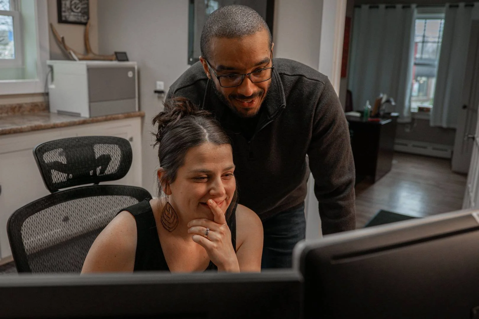 A man and woman looking at a computer monitor, with the woman appearing amused and the man leaning over her shoulder.