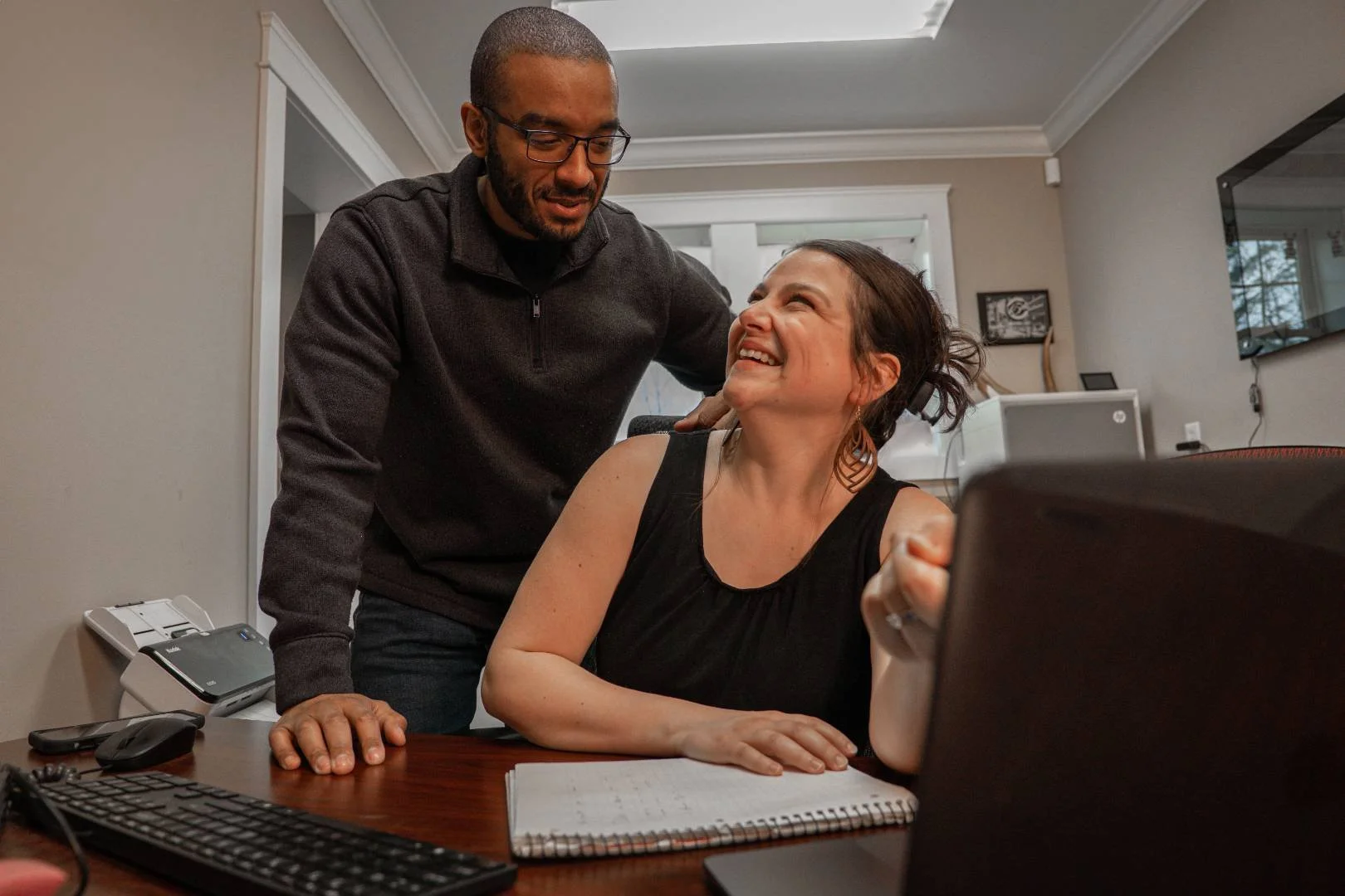 A man and a woman share a joyful moment in an office. The woman is seated, laughing and looking up at the man, who is leaning over her with a smile. The woman is wearing a black sleeveless top, and the man is dressed in a dark sweater and glasses.