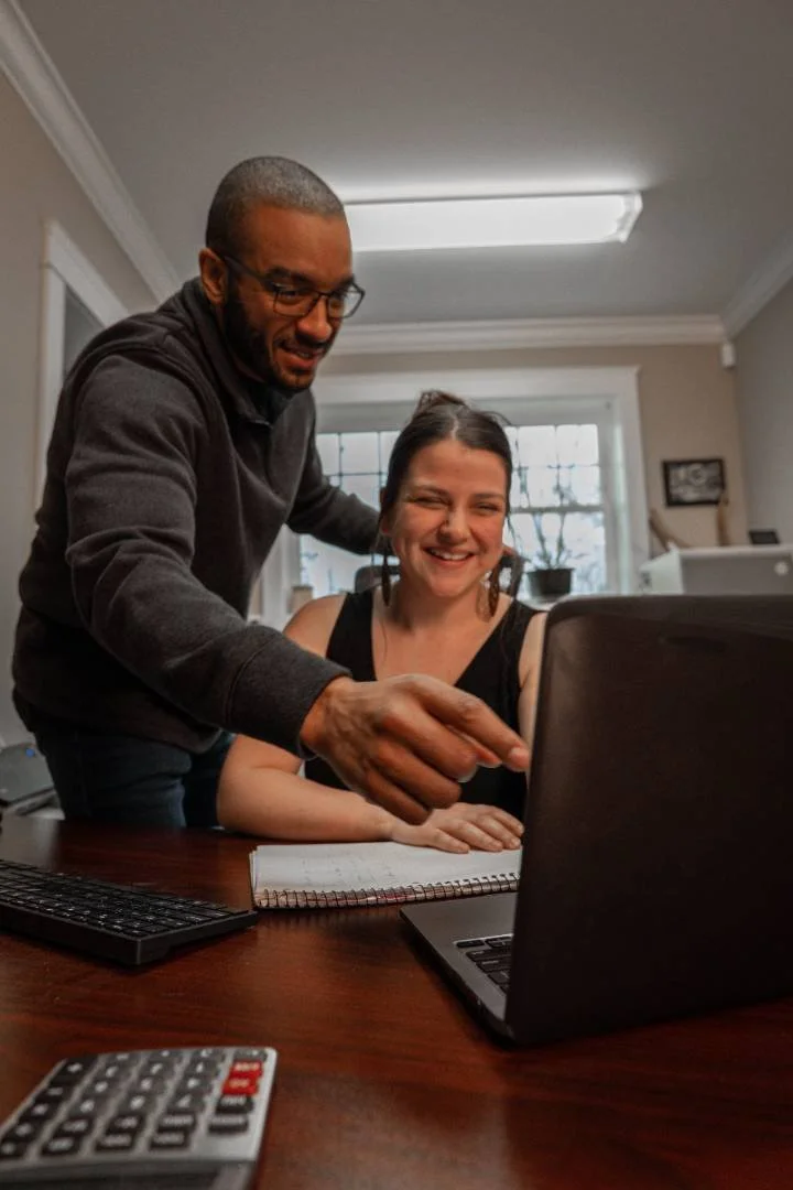 A man and a woman are looking at a laptop screen; the woman is smiling and the man appears to be explaining or pointing at something on the laptop.