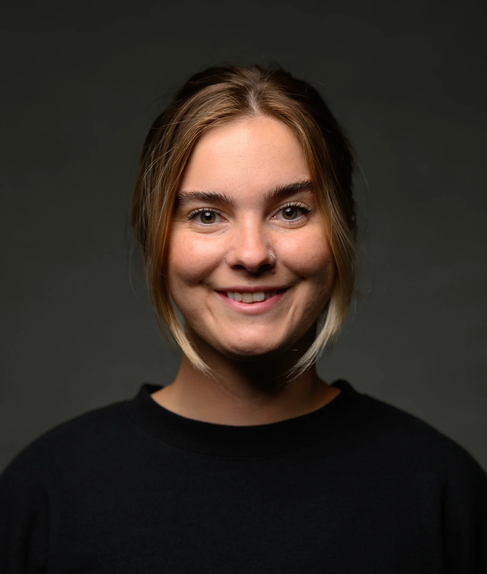 Portrait of a smiling person with light brown hair in a dark top against a dark background.