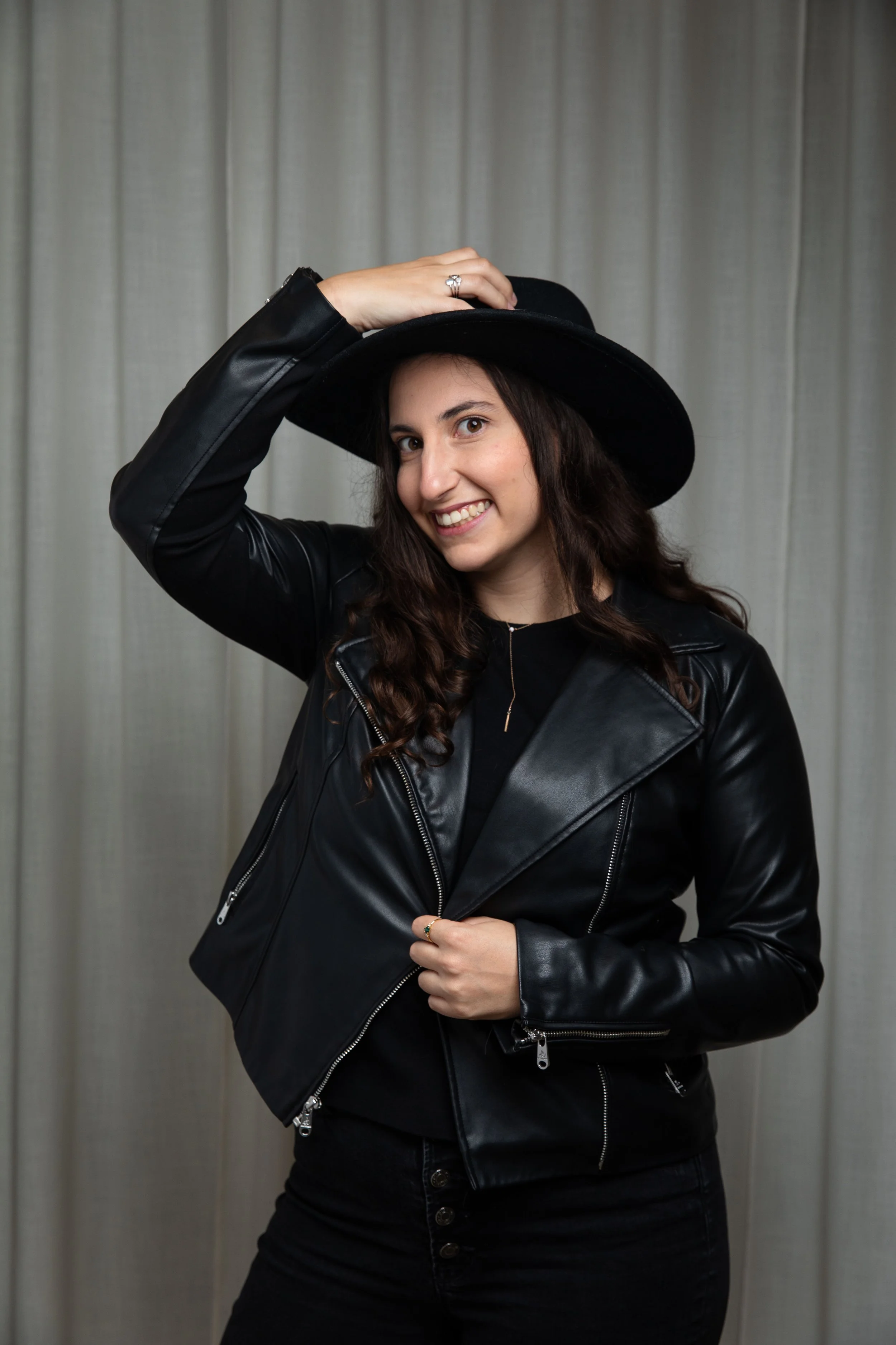 Person wearing a black leather jacket and black wide-brimmed hat, smiling and posing in front of a curtain.
