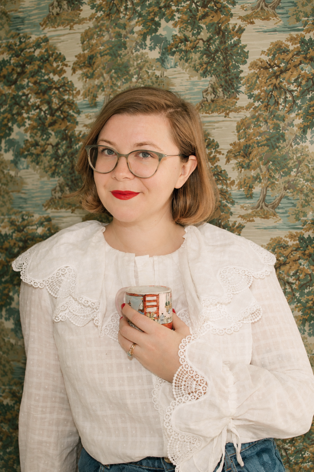 A woman with glasses and red lipstick holding a mug, standing in front of a scenic tapestry backdrop.