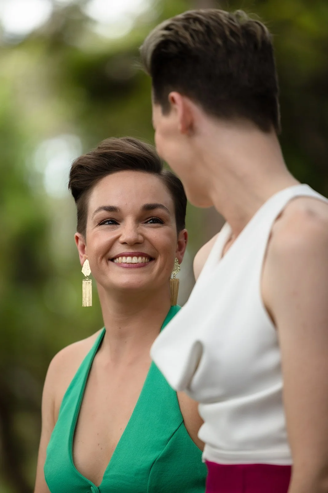 Two women are smiling and looking at each other outdoors. One woman has short hair and is wearing a green dress with tassel earrings. The other woman, whose face is partially visible, has short hair and is wearing a white sleeveless top.