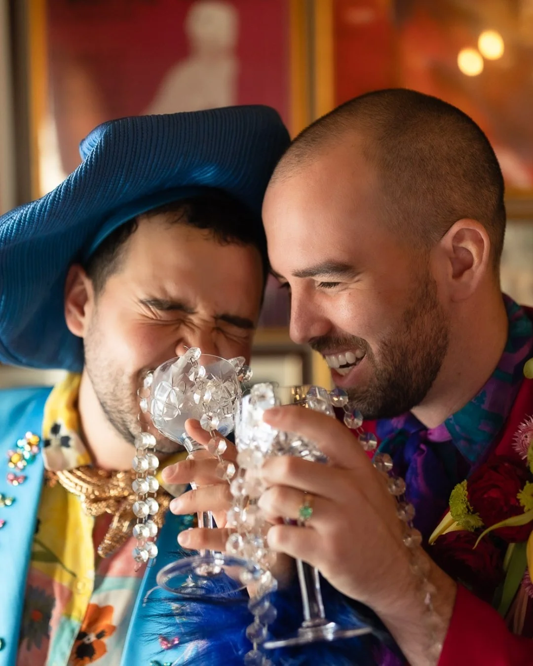 Two men smiling and close together, holding decorated glasses, in a lively, colorful setting.