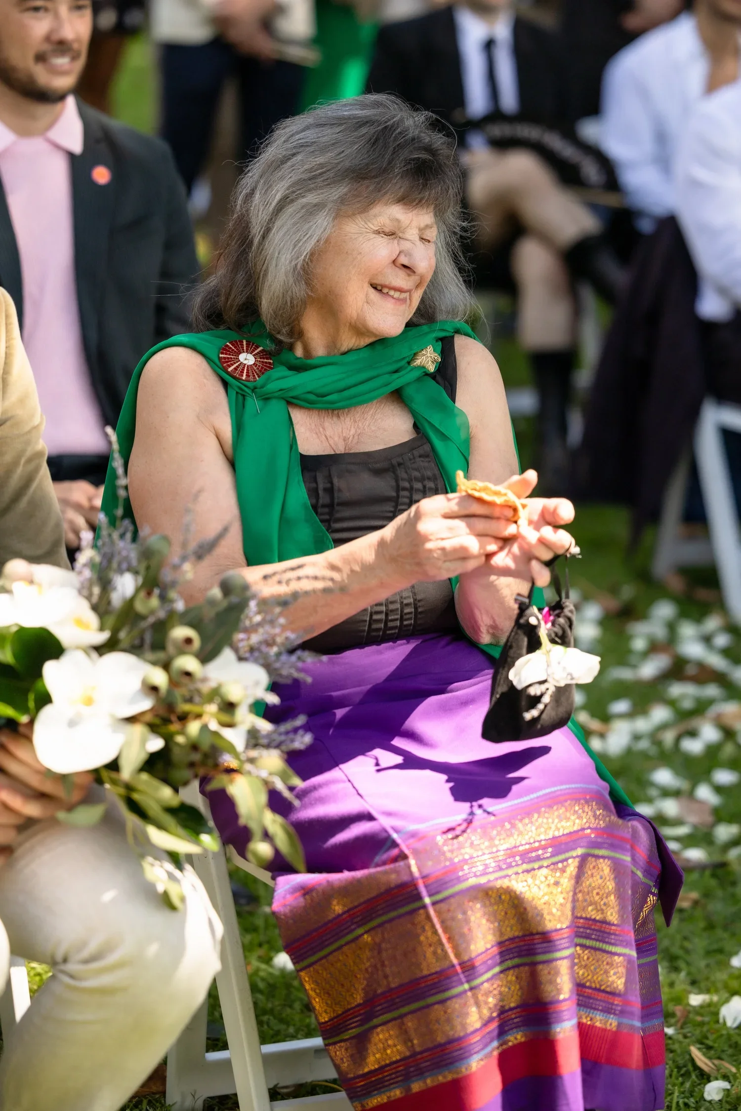 An elderly woman with grey hair smiling with eyes closed, dressed in a black top, purple and gold skirt, and a green scarf with a red pin, sitting outdoors at a wedding ceremony  in Sydney's Blackburn gardens with other people and flowers around.