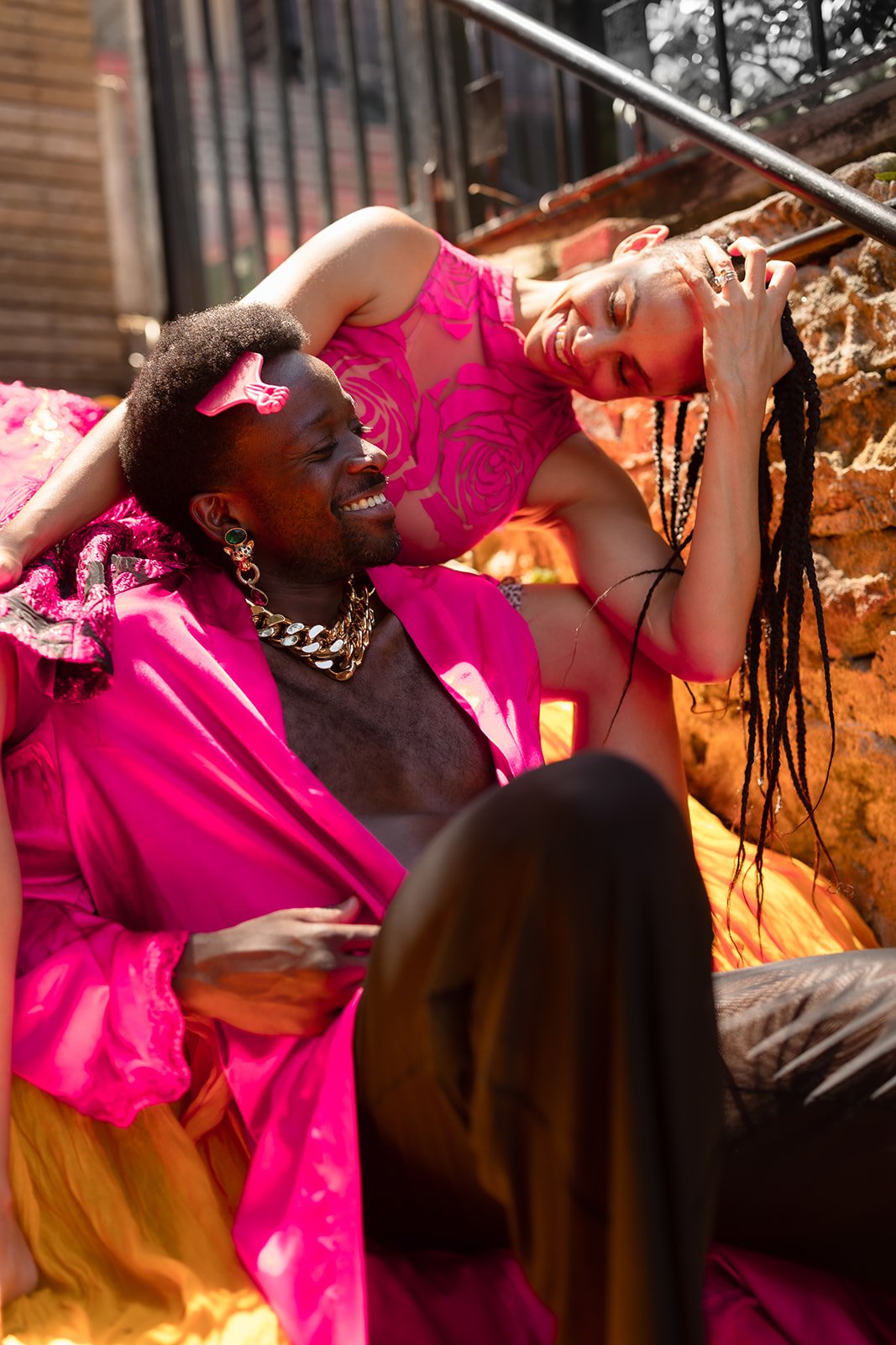 Two people, one bald woman with dreadlocks and another man with dark skin and an afro, are smiling and relaxing outdoors near a brick wall. They are dressed in vibrant pink clothing and jewelry, enjoying a joyful moment together.