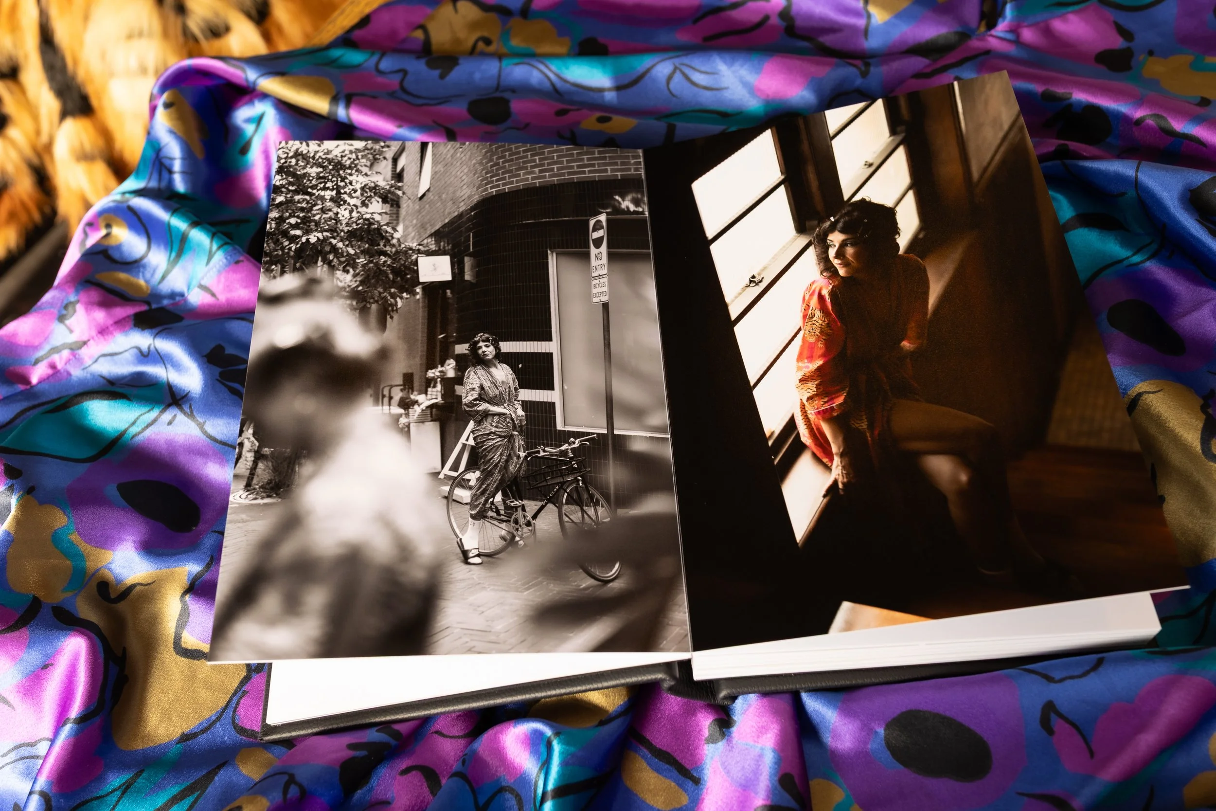 Two black and white photographs on a colorful fabric background. The left photograph shows a woman with curly hair standing with a bicycle on a city street, with trees and a modern building in the background. The right photograph shows a woman with curly hair sitting by a window, looking to her left, in an indoor setting.