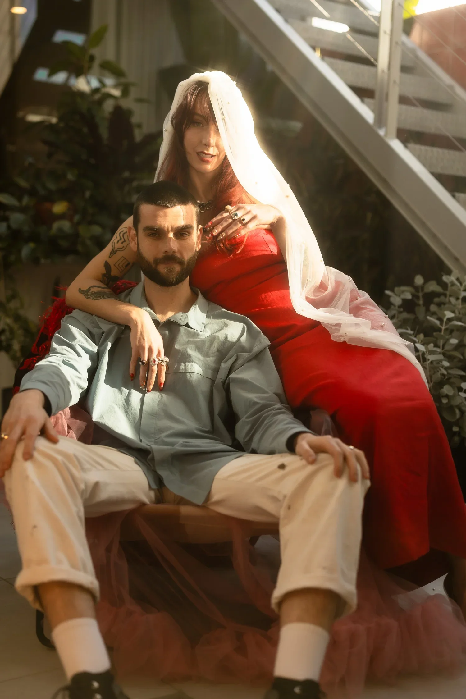 A man with a beard in beige pants and a light grey shirt is sitting on a chair, with a woman with red hair, tattoos, and wearing a red dress and a white veil sitting behind him, her arm resting on his shoulder. They are in an indoor setting with plants and a staircase in the background.