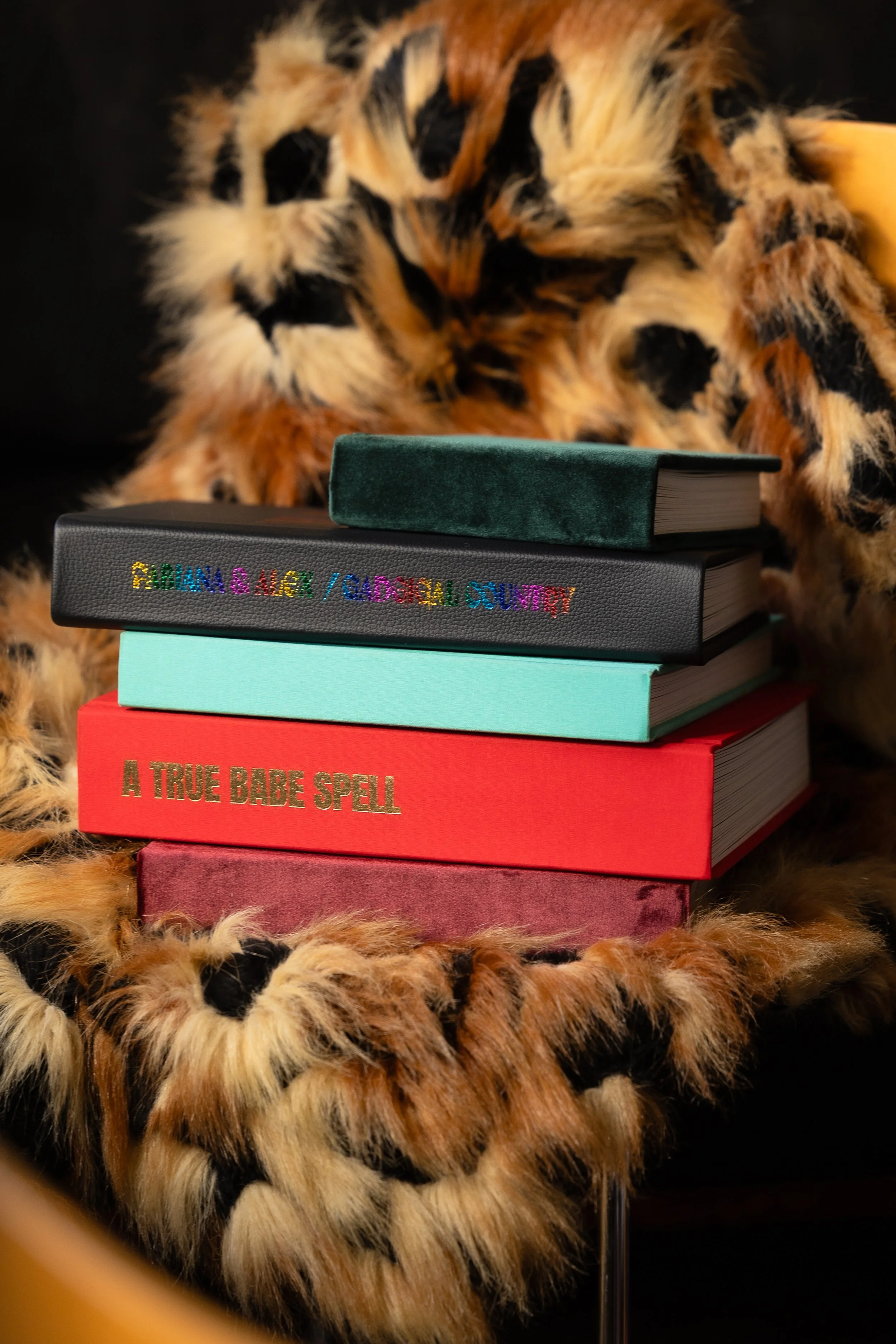Stack of five colorful books with titles: "GIRL GANG & GENDER / GENDER CONFLICT" (black cover), a green book, a light blue book, "A TRUE BABE SPELL" (red cover), and a purple book, all on a furry animal patterned surface with a dark background.