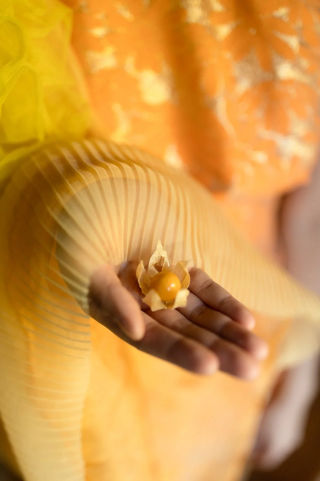 A person holding a yellow lotus flower-shaped arrangement with a small round yellow object in the center, dressed in a yellow and orange patterned garment.