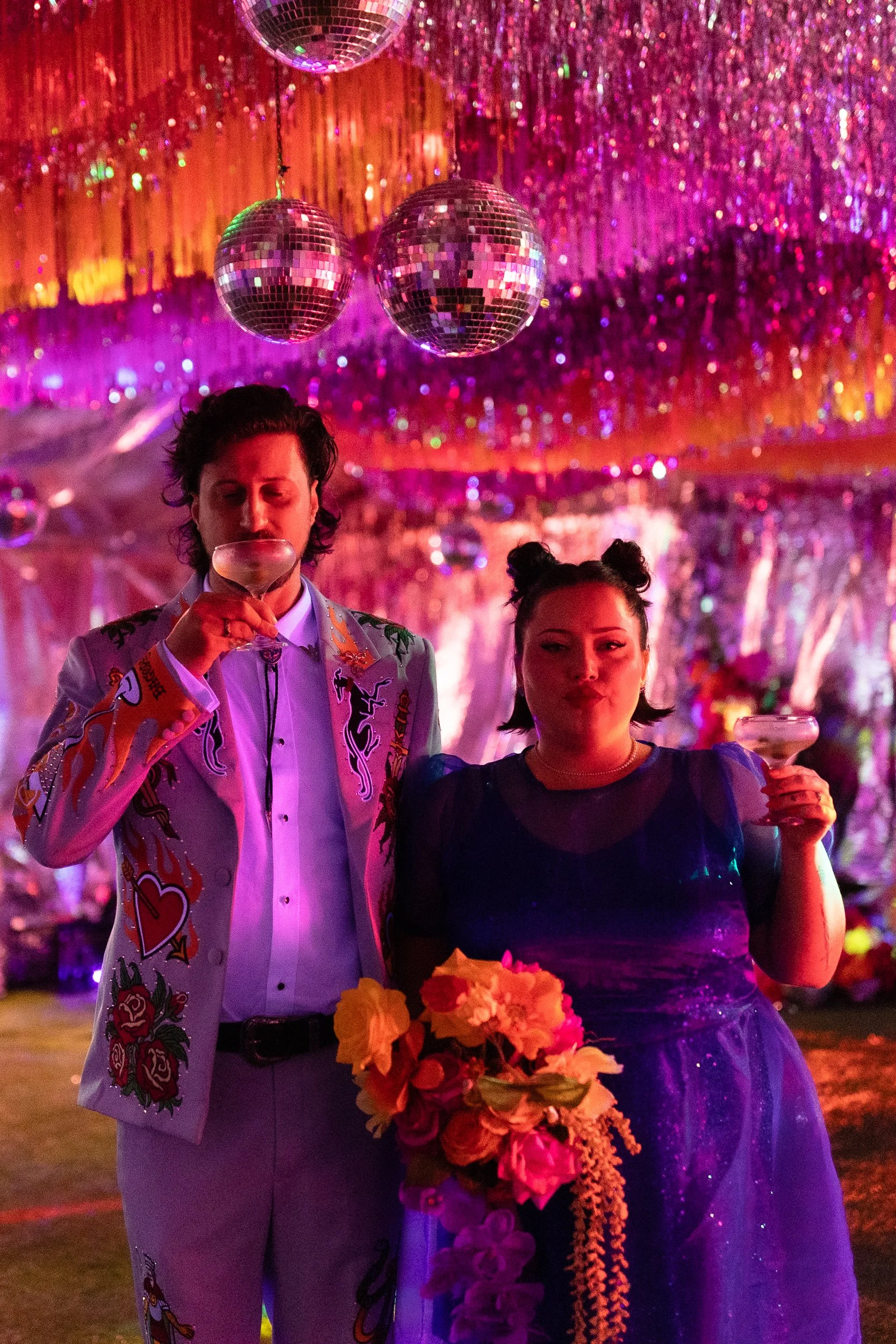 Two people celebrating at a party with disco balls and vibrant pink, purple, and orange decorations. One person is wearing a colorful jacket with various designs and holding a drink, while the other is wearing a dark dress and holding a flower bouquet.