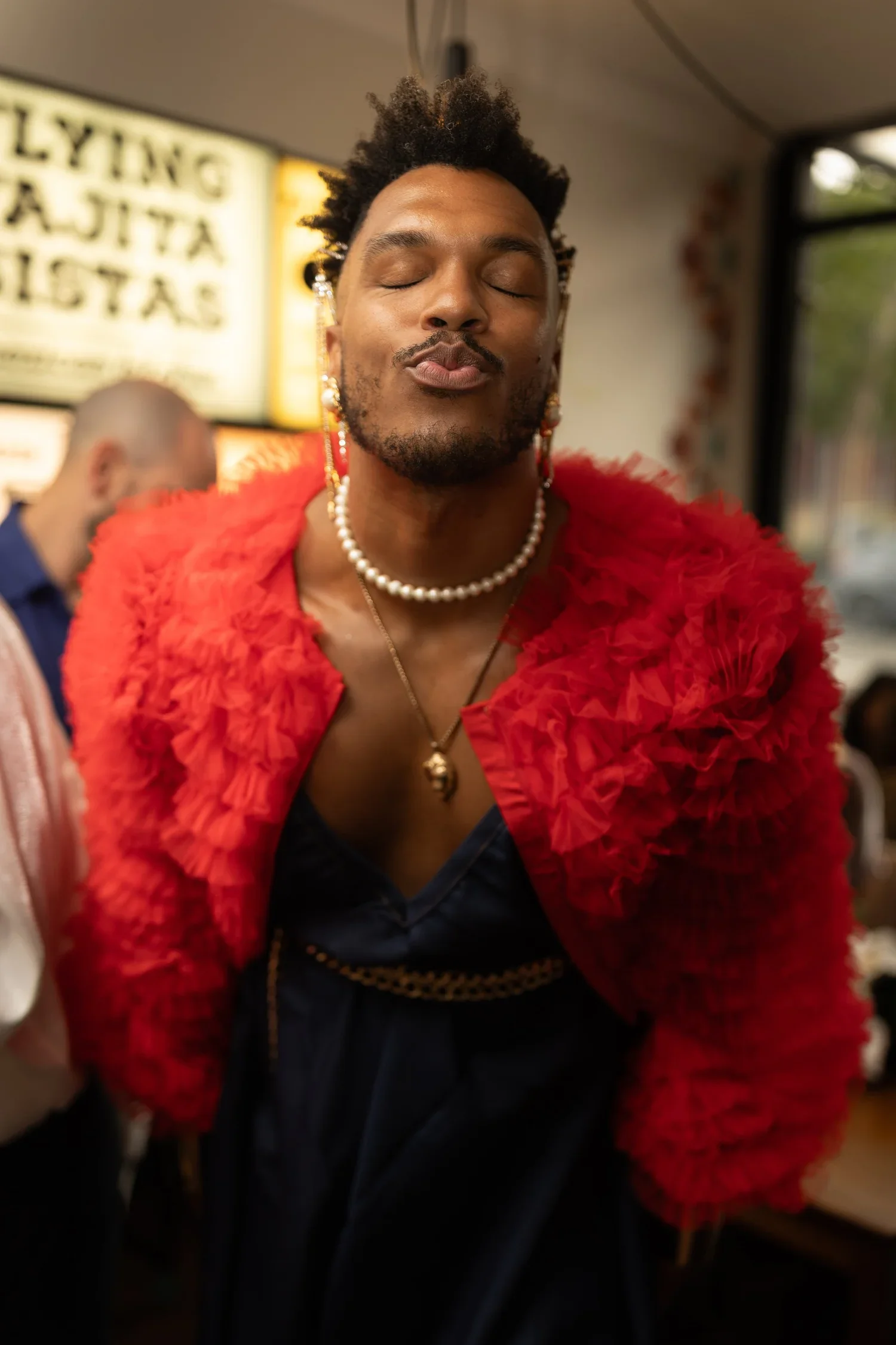 Person with closed eyes, wearing a red textured jacket, pearl necklace, and earrings, inside a building with blurry background for a wedding reception in Sydney's Inner West