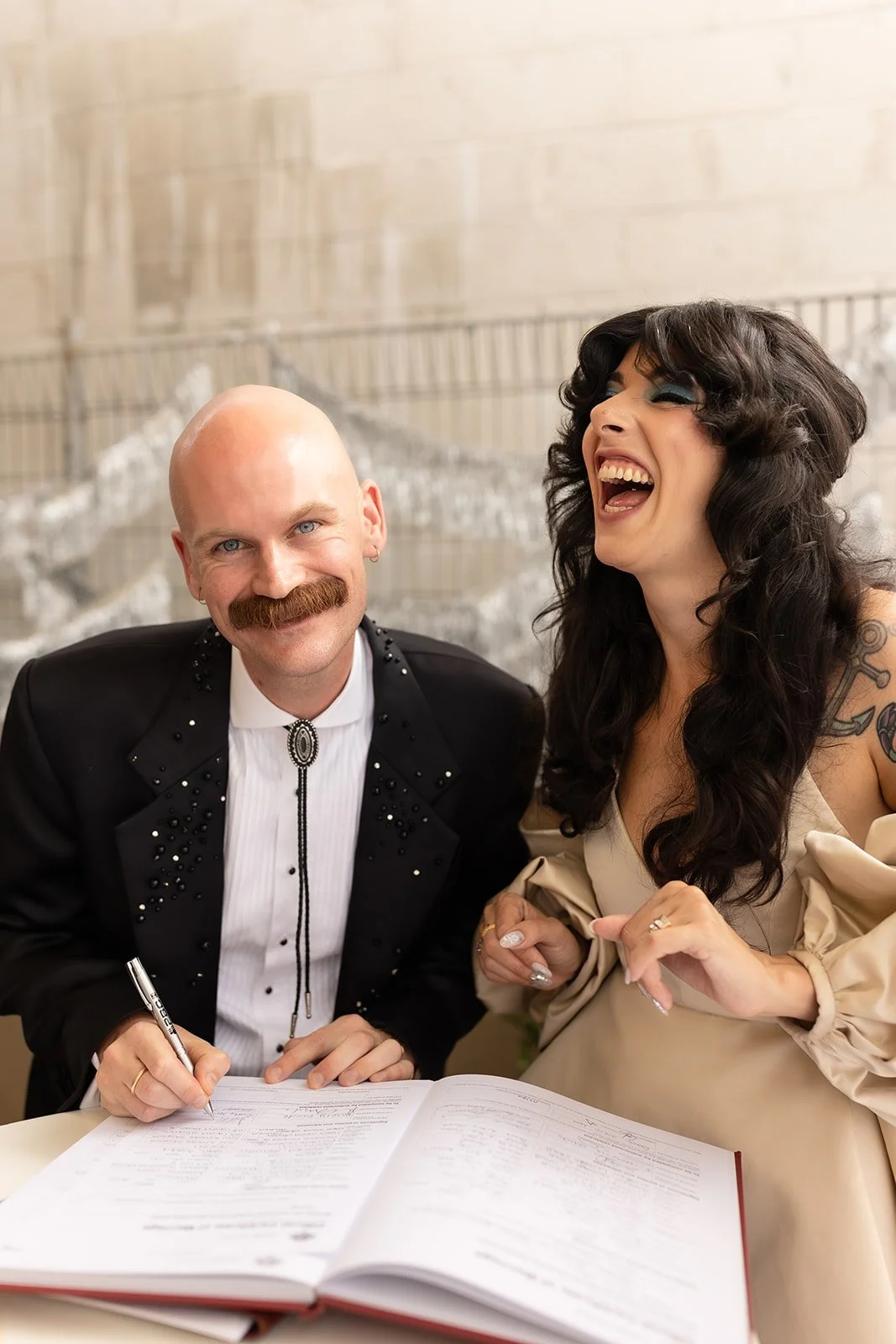 A man with a bald head and mustache and a woman with long dark hair and tattoos laughing together while signing a document at a table.