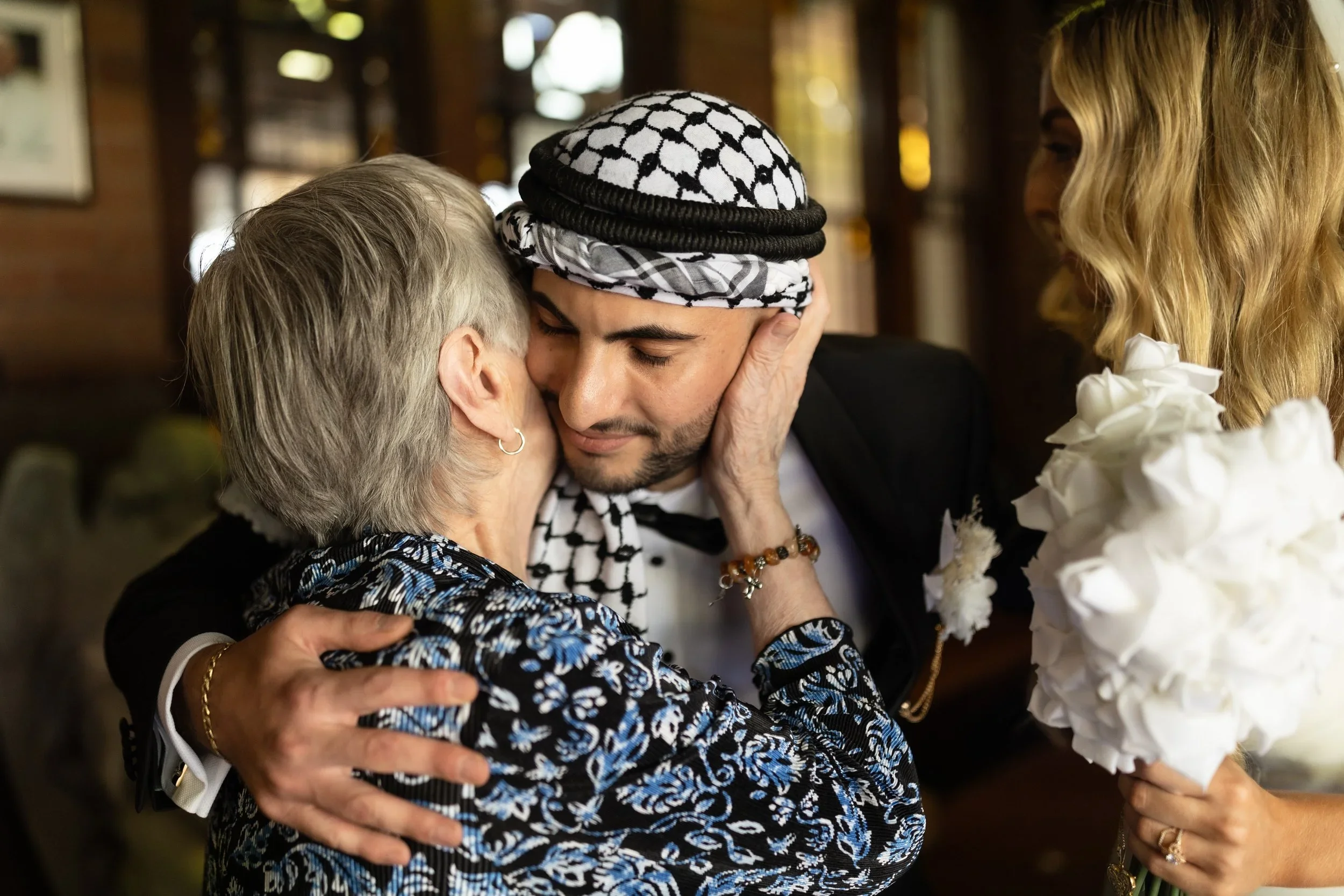 A young man with a black and white keffiyeh headscarf hugging an elderly woman, both with eyes closed, in an embrace in a warmly lit indoor setting. Another woman with blonde hair is visible on the right, holding white flowers.