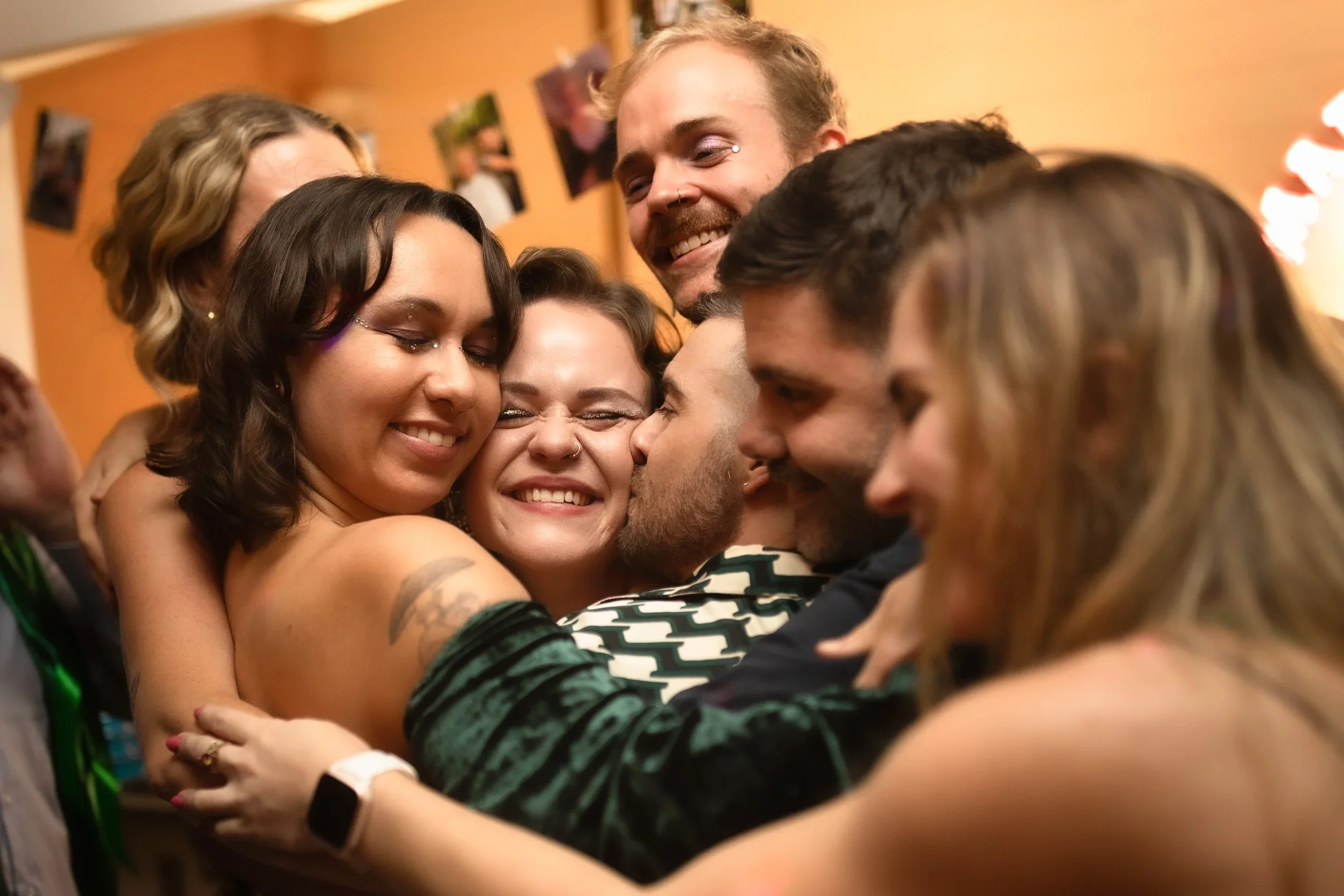 A group of diverse friends are hugging each other closely and smiling joyfully at a party or gathering indoors. The background is blurry with warm lighting and wall decorations.