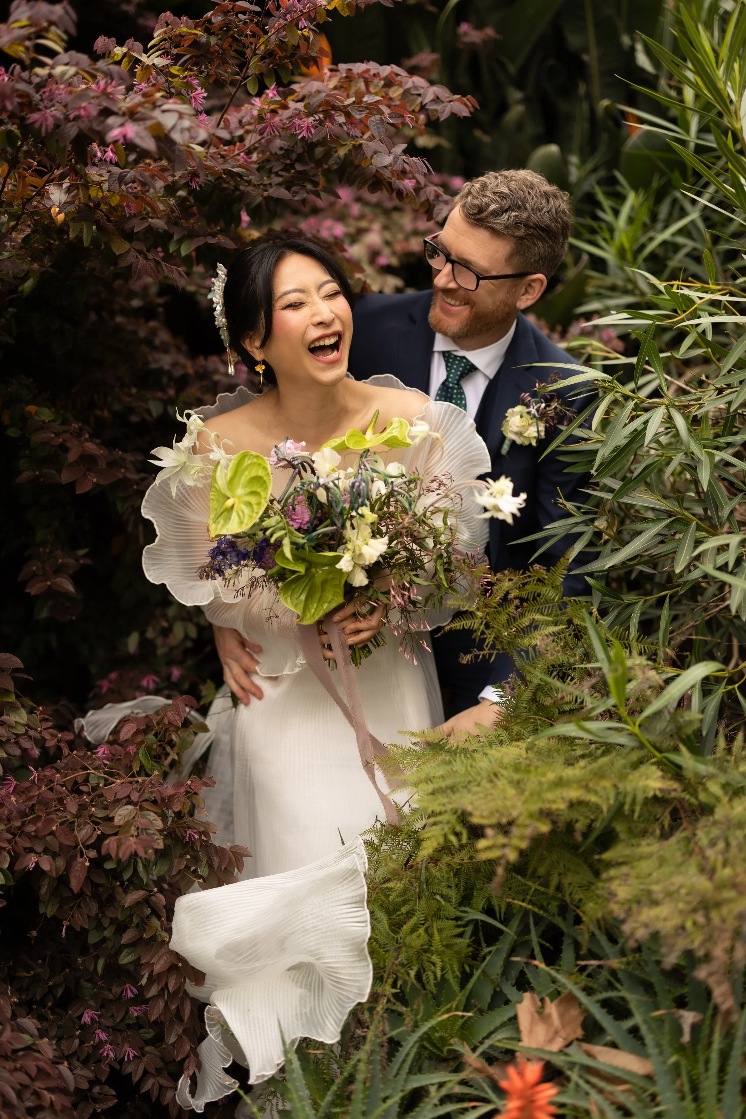 A couple in wedding attire sharing a joyful moment in a lush garden surrounded by various green and purple plants.