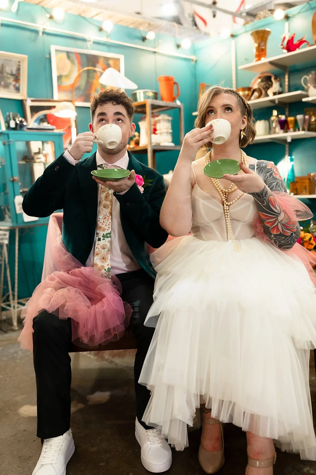 A man and woman dressed in wedding attire, sitting in a colorful vintage shop, drinking from white cups, with pink tulle skirts under their suits, surrounded by shelves of ceramics and decorative items.