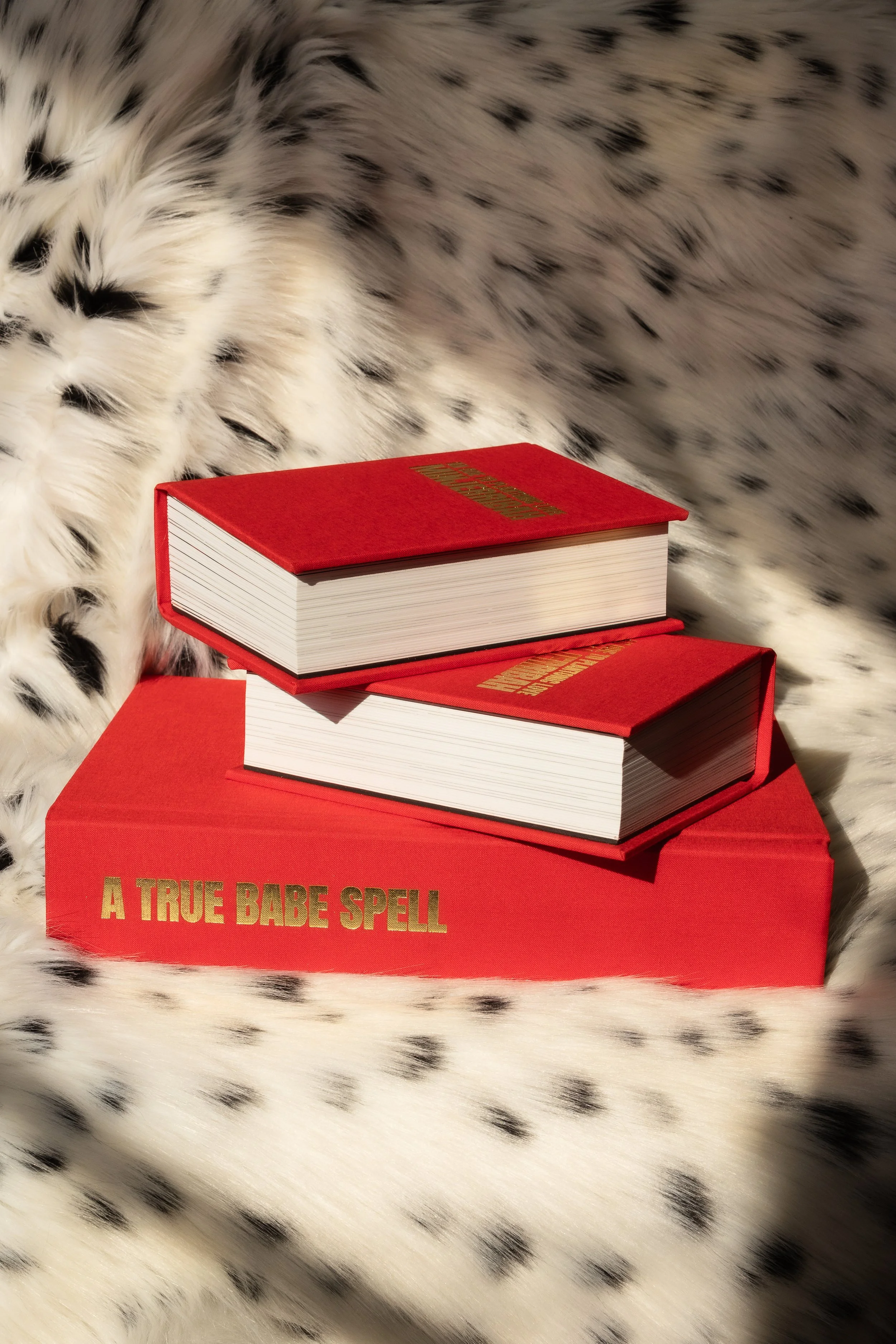 Three red books with gold lettering are stacked on top of a red box that says "A TRUE BABE SPELL" in gold letters. The background is fuzzy white fur with black spots.