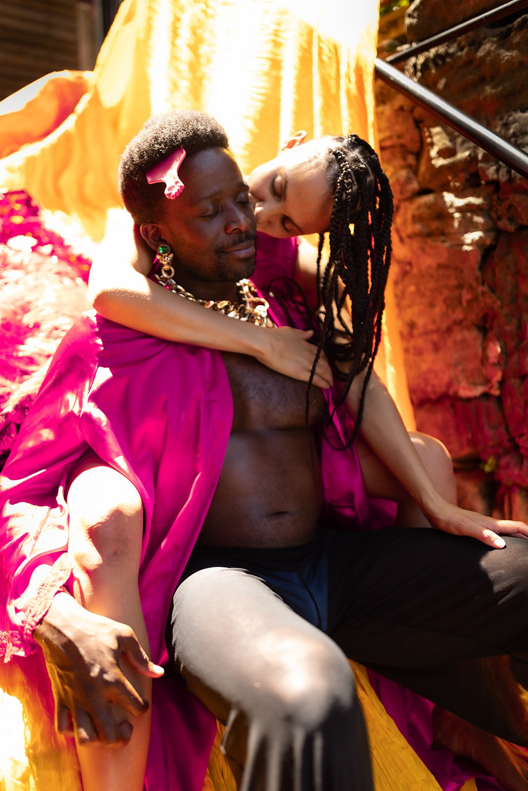 A man and woman embrace on a colorful outdoor backdrop with yellow fabric and red rocks, showing an intimate moment.