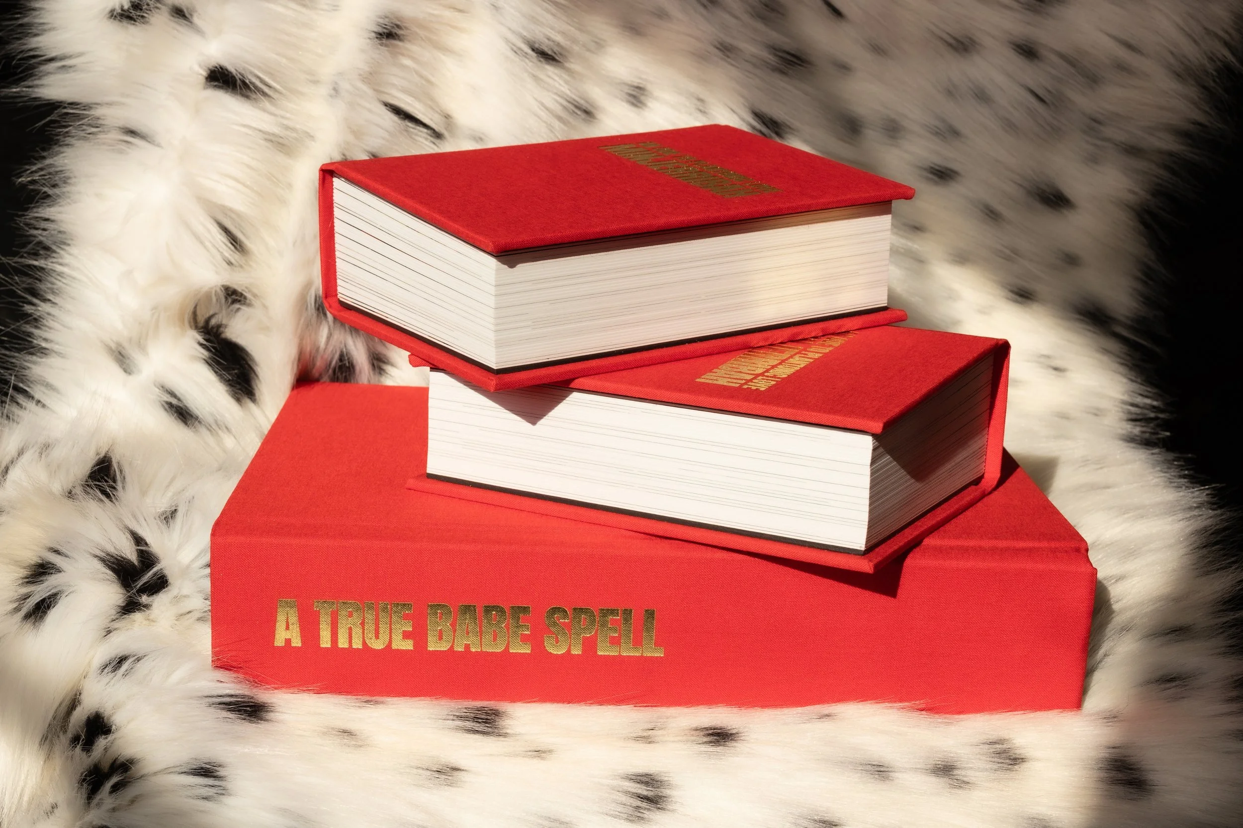 Three red hardcover books stacked on top of each other on a red box with a faux animal fur background. The top book has gold text on the cover, and the bottom box has gold text reading 'A TRUE BABE SPELL'.