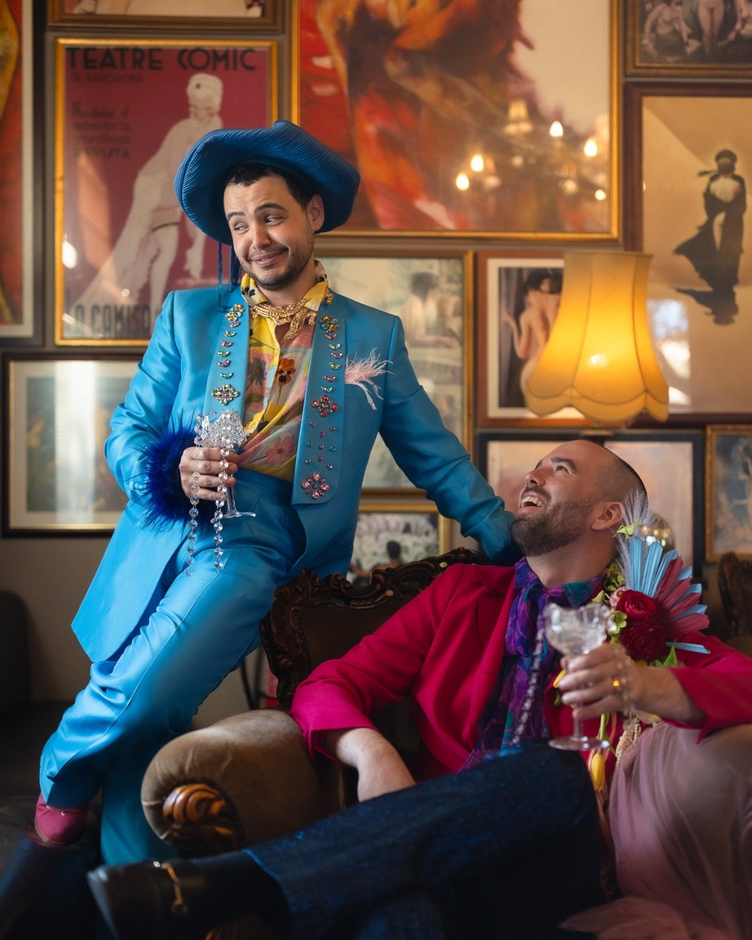 Two men in colorful, flamboyant outfits, one standing and one sitting, in a decorated room with framed art and warm lighting, enjoying a conversation with drinks.