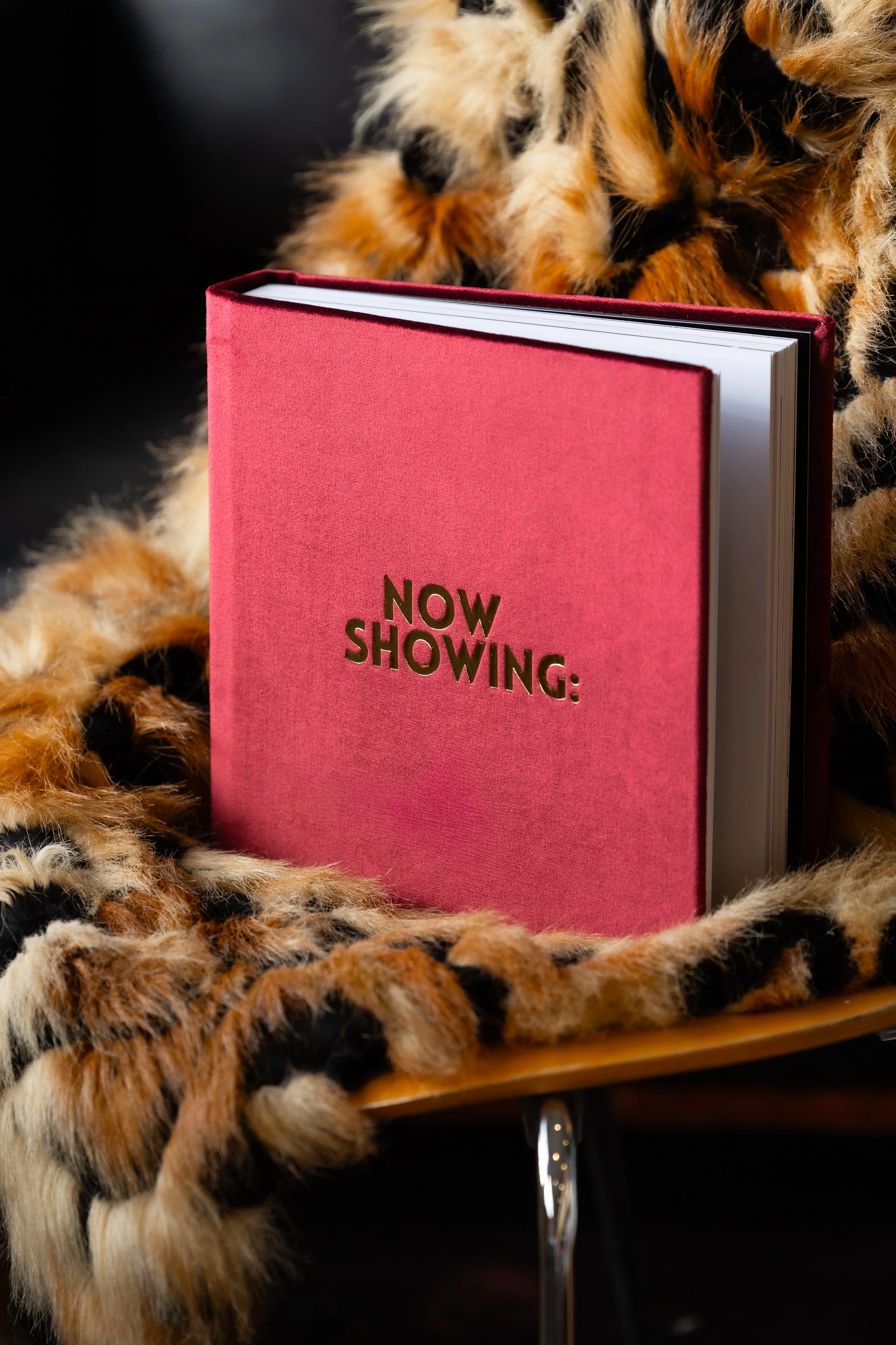 A cheetah lying on a table with a red book titled 'Now Showing:' in front of it, surrounded by a leopard-print fabric.