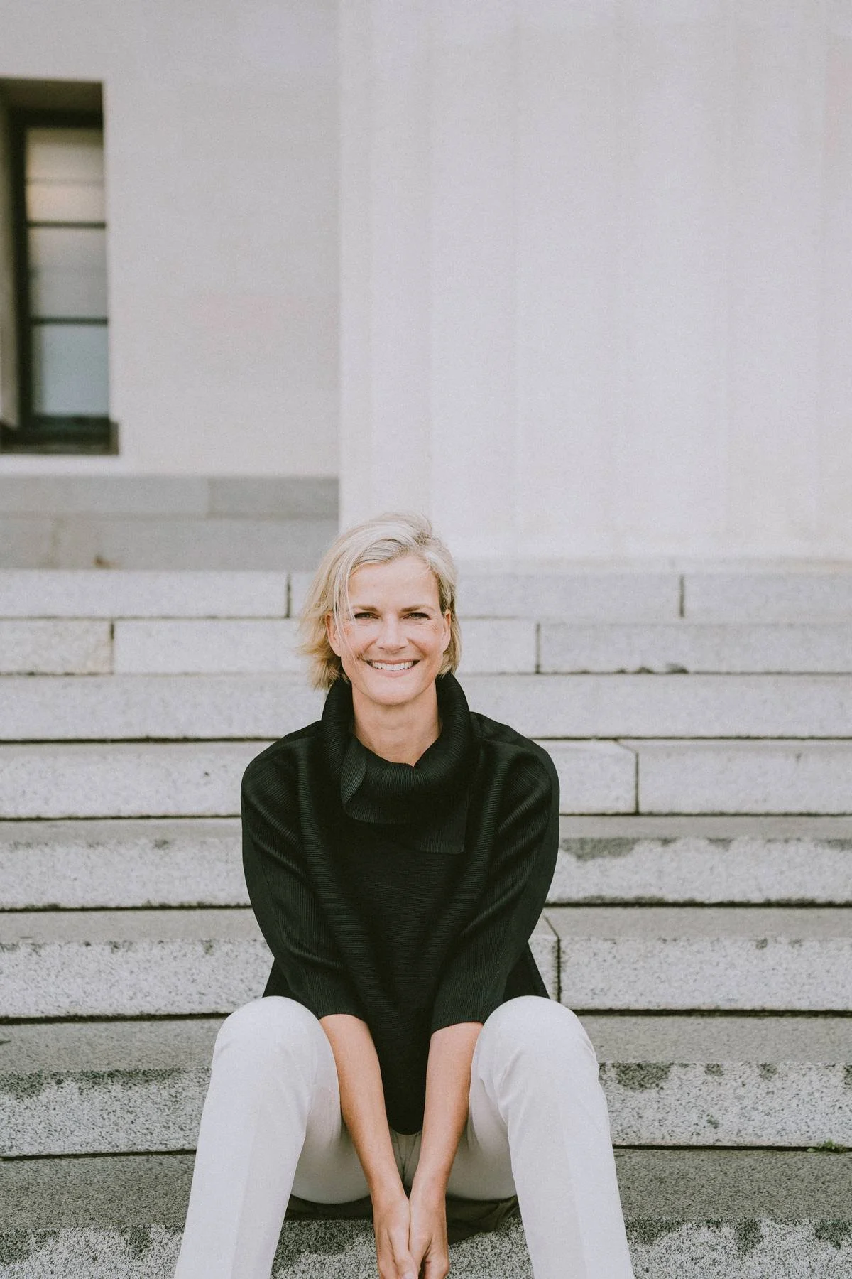 A woman with short blonde hair sitting on steps outside a building, smiling at the camera. She is wearing a black top and light-colored pants.