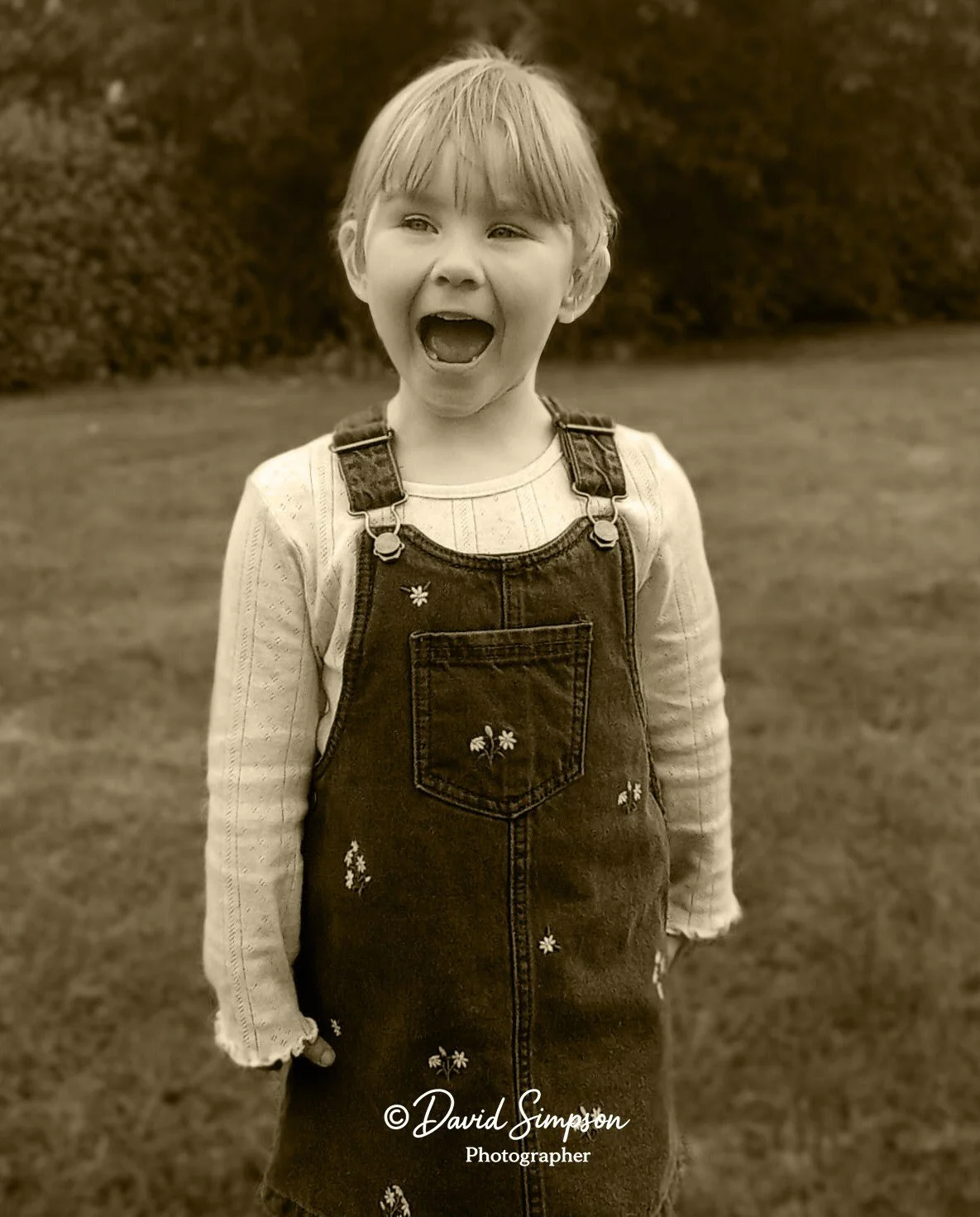 A young girl with short hair, wearing a long-sleeve shirt and a dress with embroidered flowers, standing outdoors on a grassy field with trees in the background, with her mouth open as if shouting or laughing.