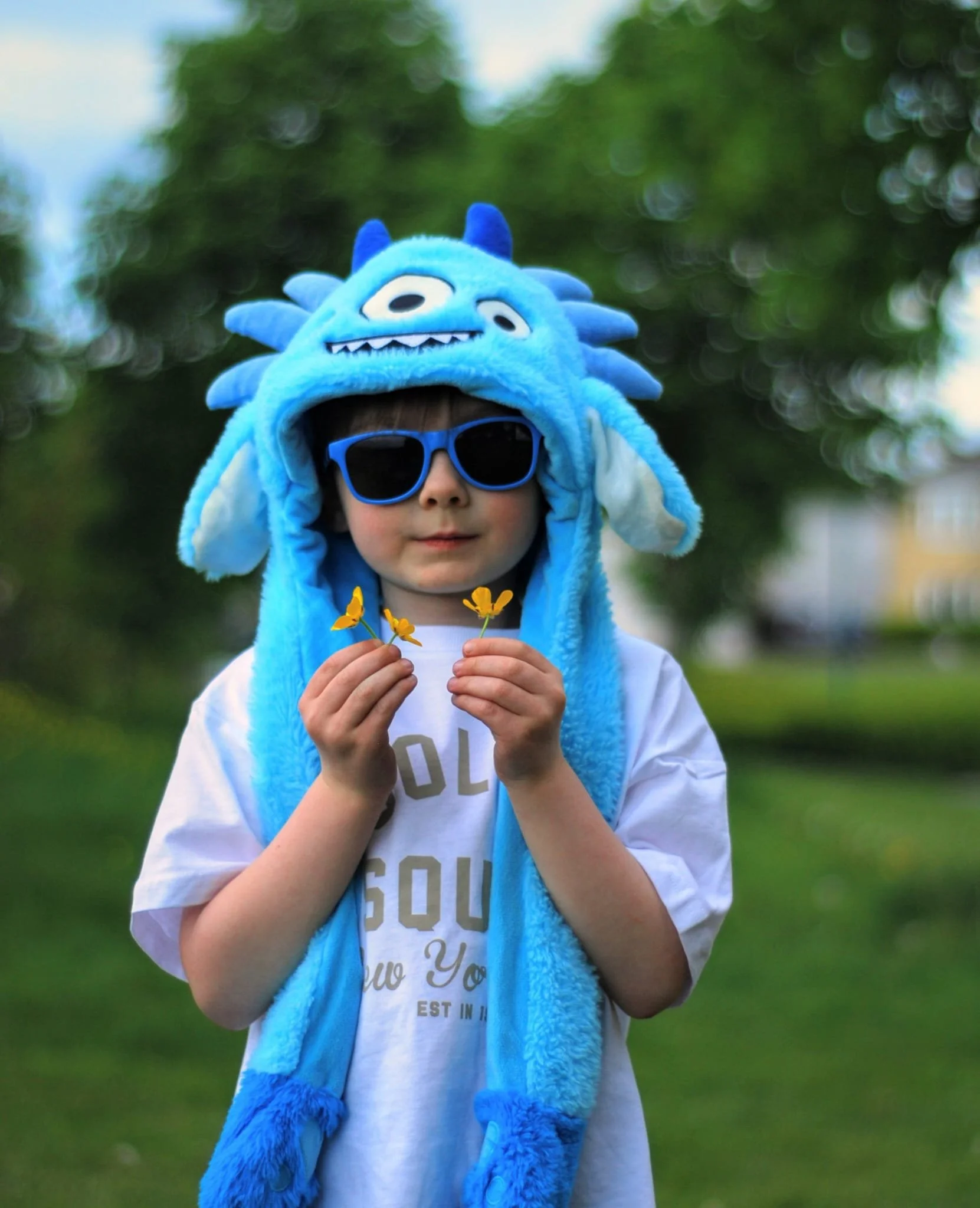 A young boy wears a blue monster-themed hat with multiple facial features, blue sunglasses, and a white T-shirt. He holds yellow flowers in his hands and stands outdoors in a grassy area with trees in the background.
