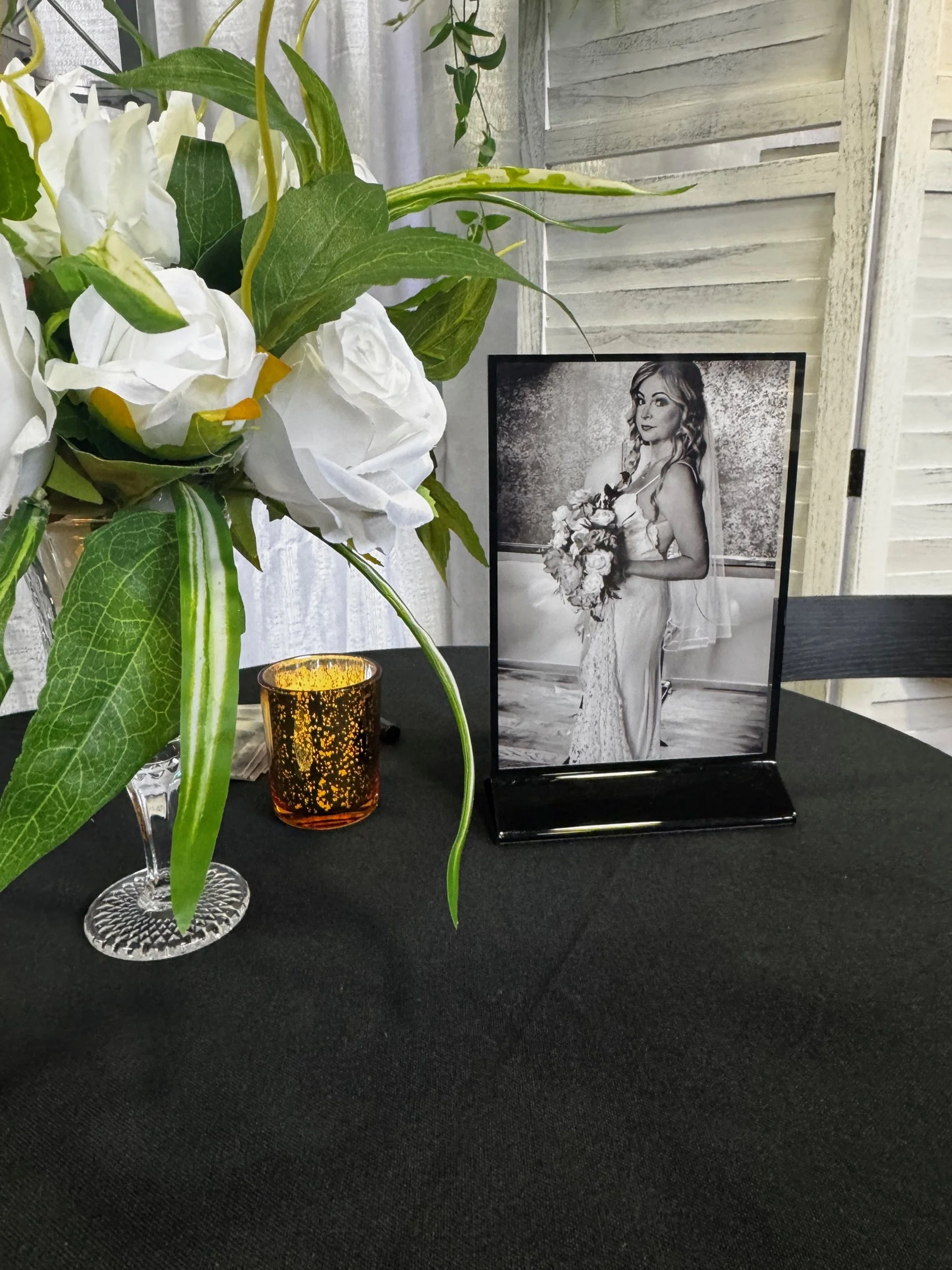 A wedding table decoration with a black-and-white photo of a bride holding a bouquet, a floral arrangement with white flowers and green leaves, and a small lit candle in a decorative holder on a black tablecloth.