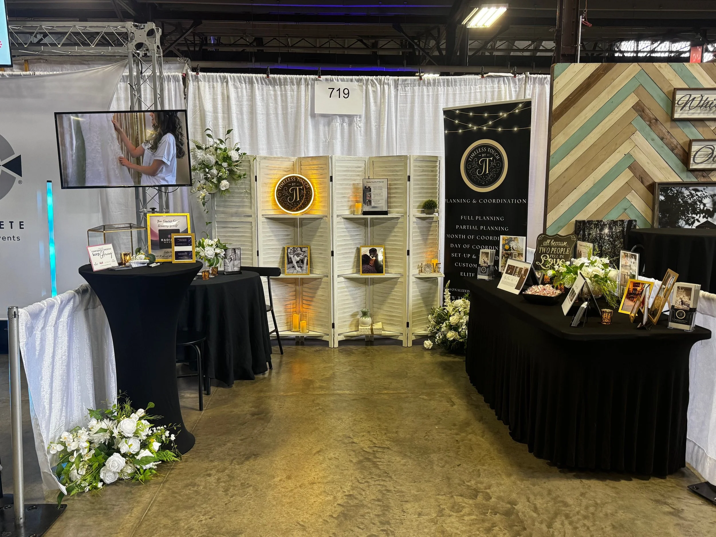 Exhibition booth for a planning and coordination service decorated with white flowers, framed pictures, and candles, with a black tablecloth and information signs, set in an indoor event space.