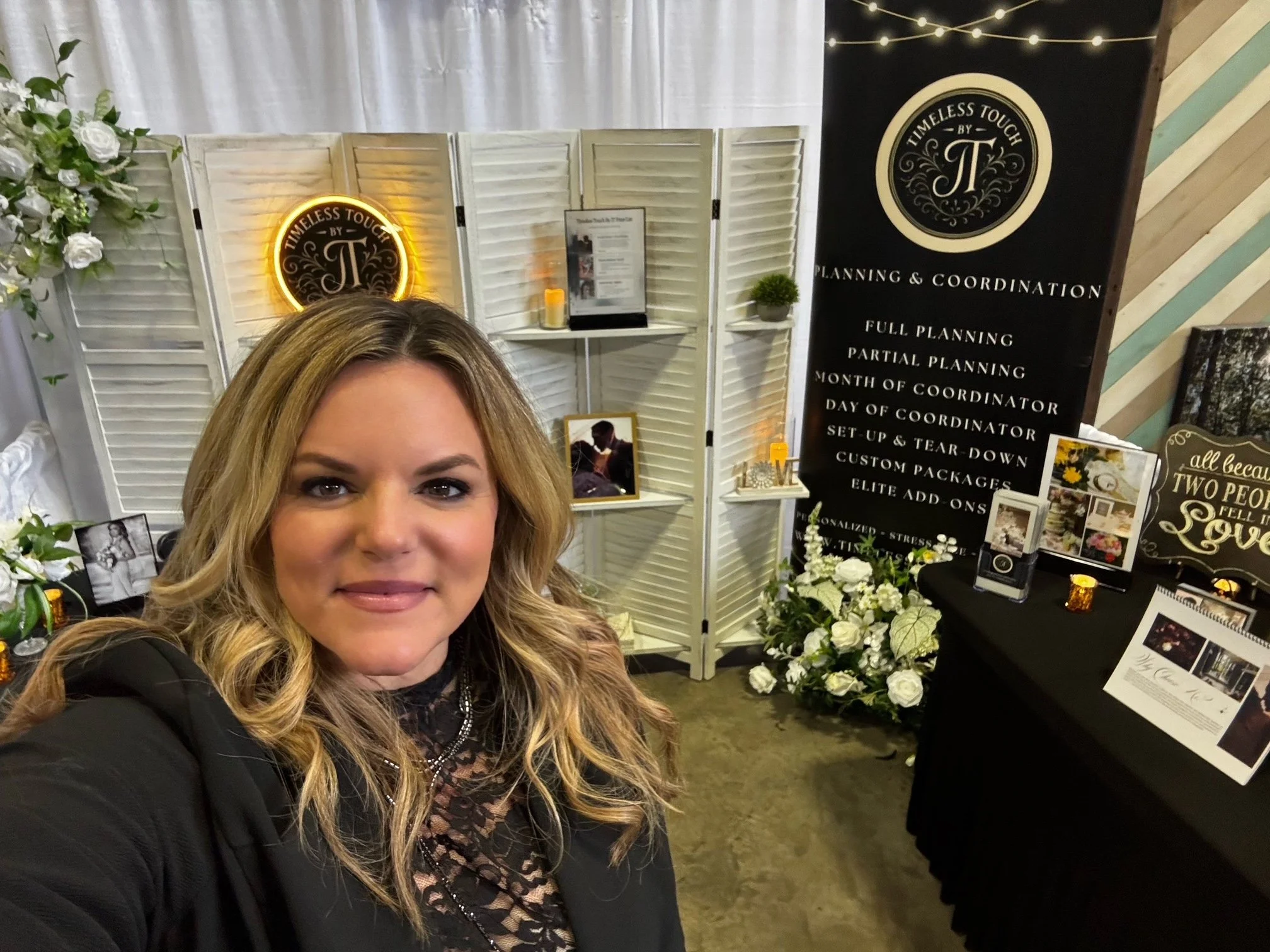 A woman with wavy blonde hair smiling for a selfie at an event booth. The booth features a tall black sign with gold and white lettering, white folding screens, and floral arrangements with white roses. The sign lists planning and coordination services, including full, partial, month, and day-of planning, plus custom packages and add-ons.