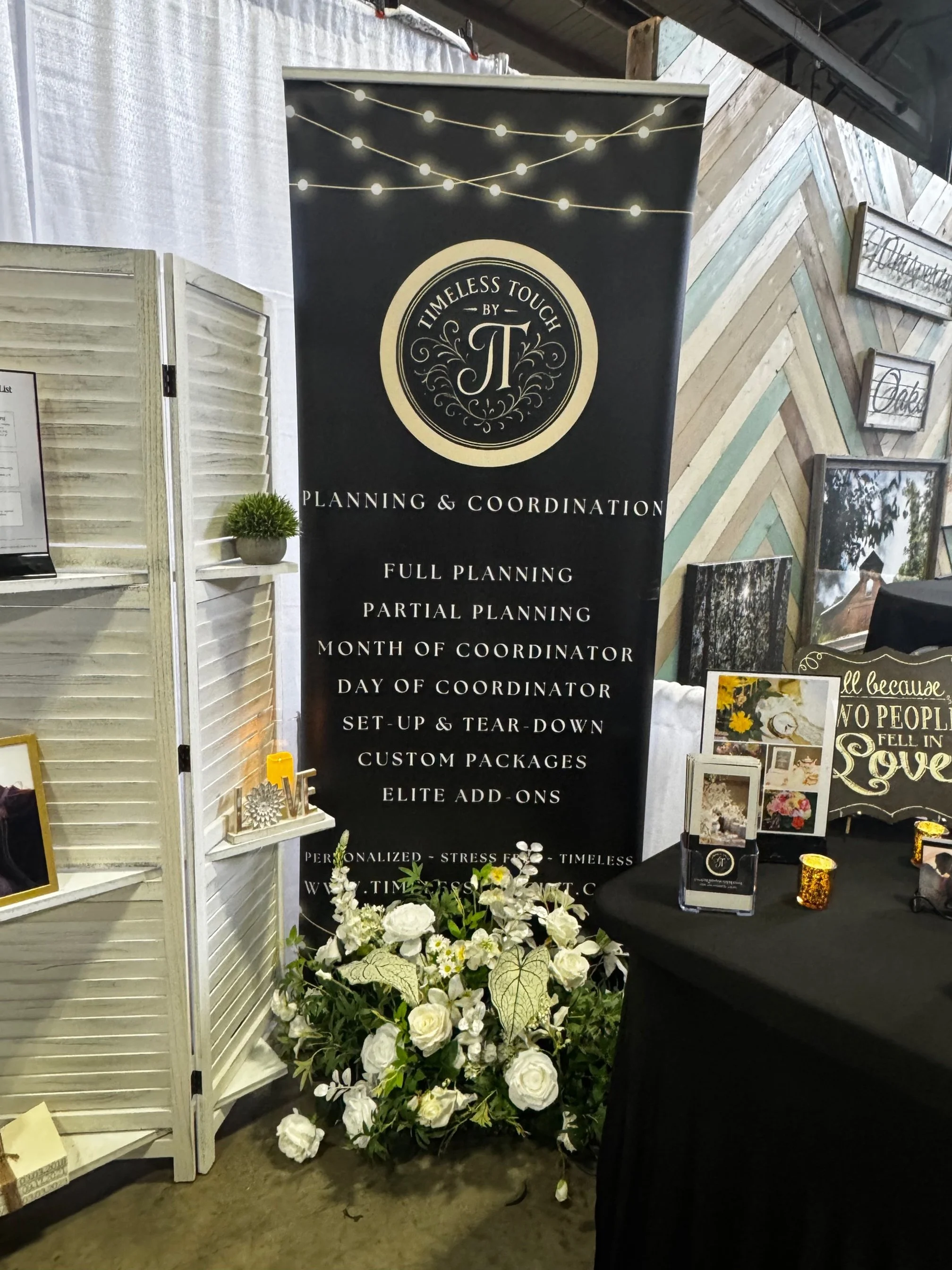 A tall black banner displays services like planning and coordination for events, with decorative string lights at the top, and is surrounded by white flowers at the base and small decorative items to the right.