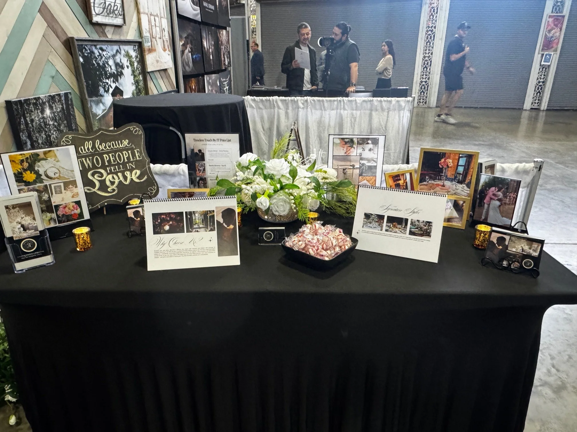 Table display at a wedding or event with framed photos, signs, and flowers, including a sign that reads 'all because TWO PEOPLE FELL IN Love'.