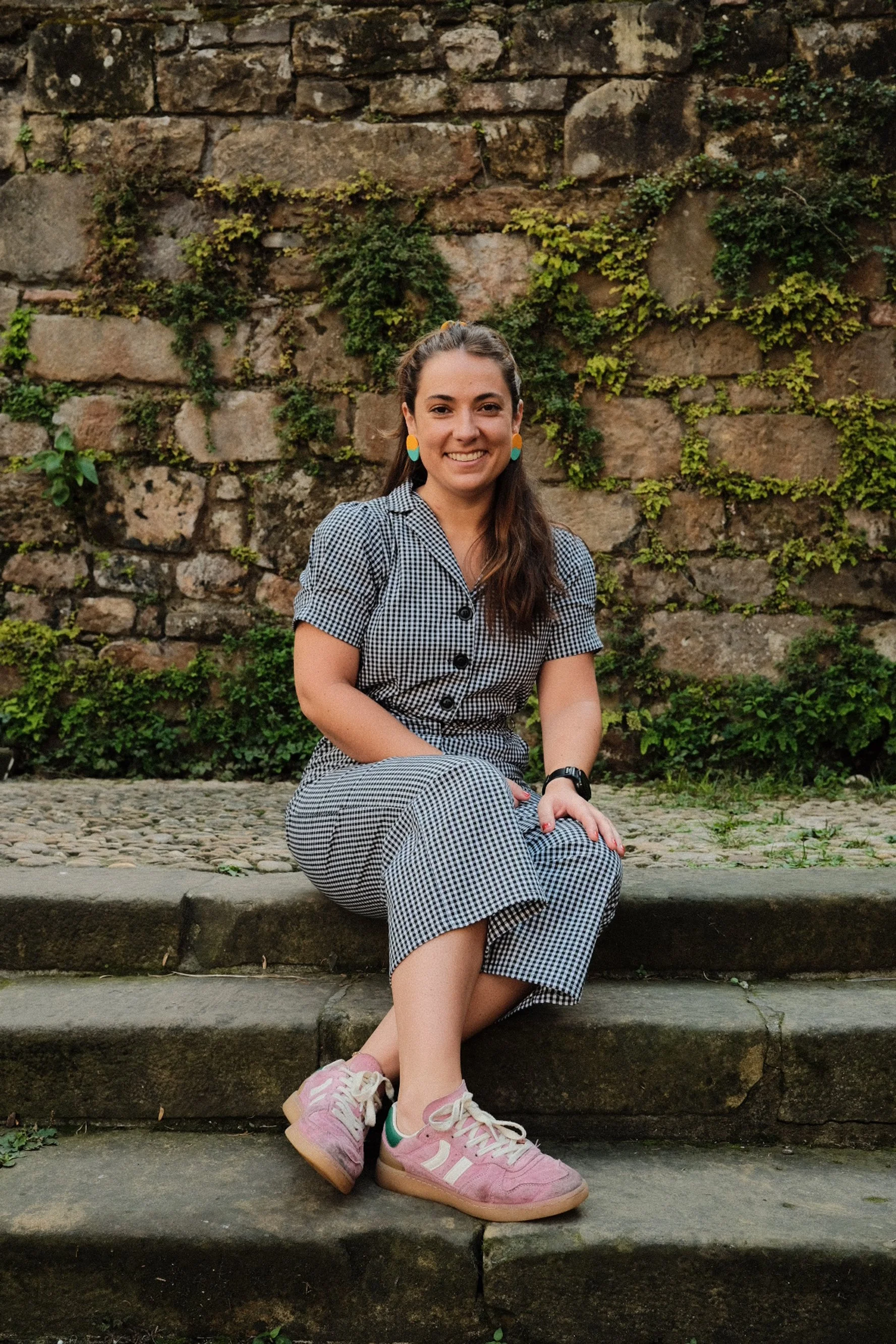 Person with topknot hairstyle wearing colorful earrings against a green background.