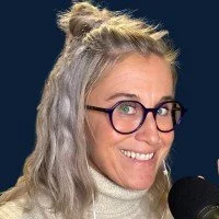 A woman with blonde, curly hair styled in a half-up bun, wearing glasses and a light-colored turtleneck sweater, smiling while speaking into a microphone.