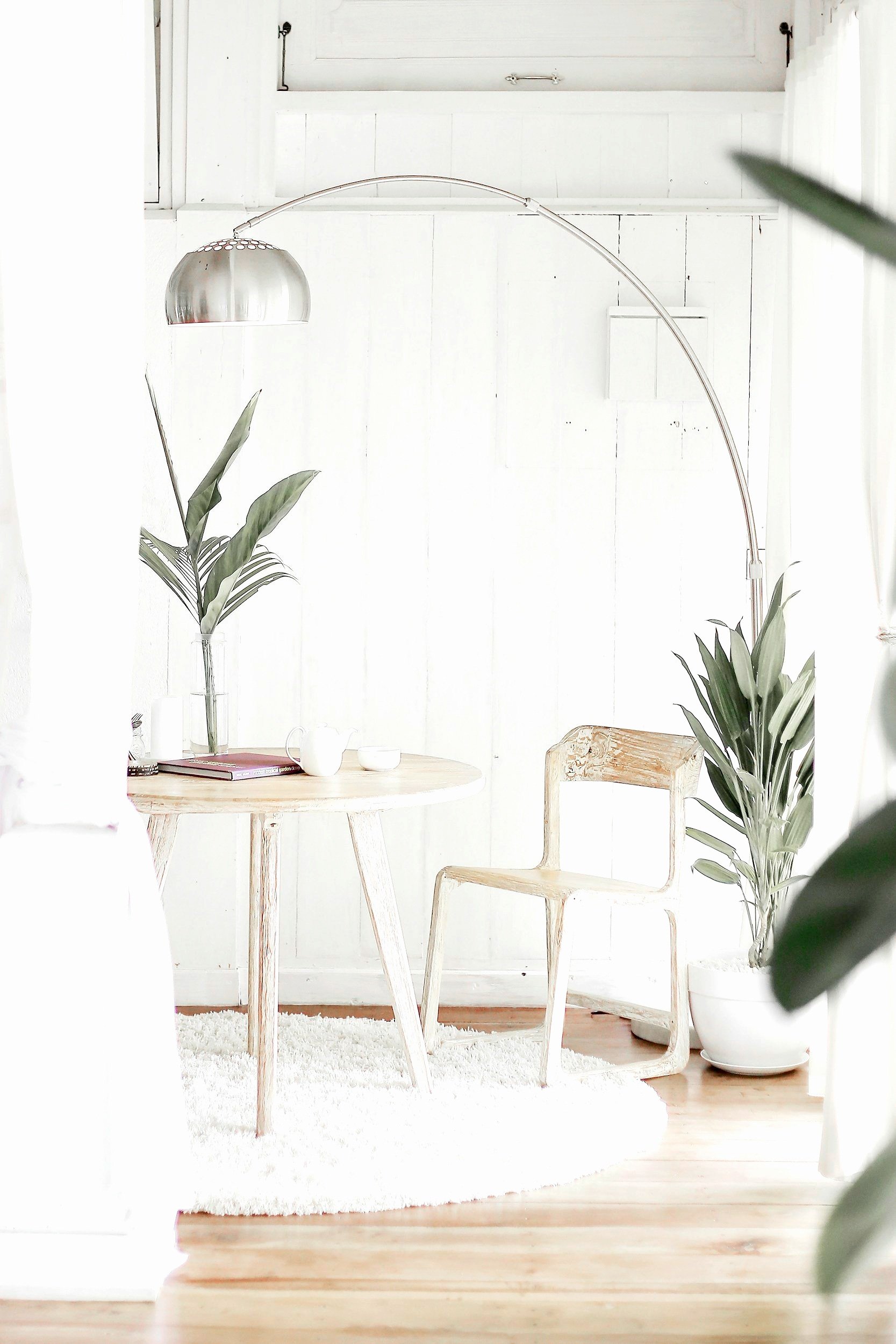 Minimalist white room with a wooden table, curved metal floor lamp, wooden chair, potted plants, and decorative items.