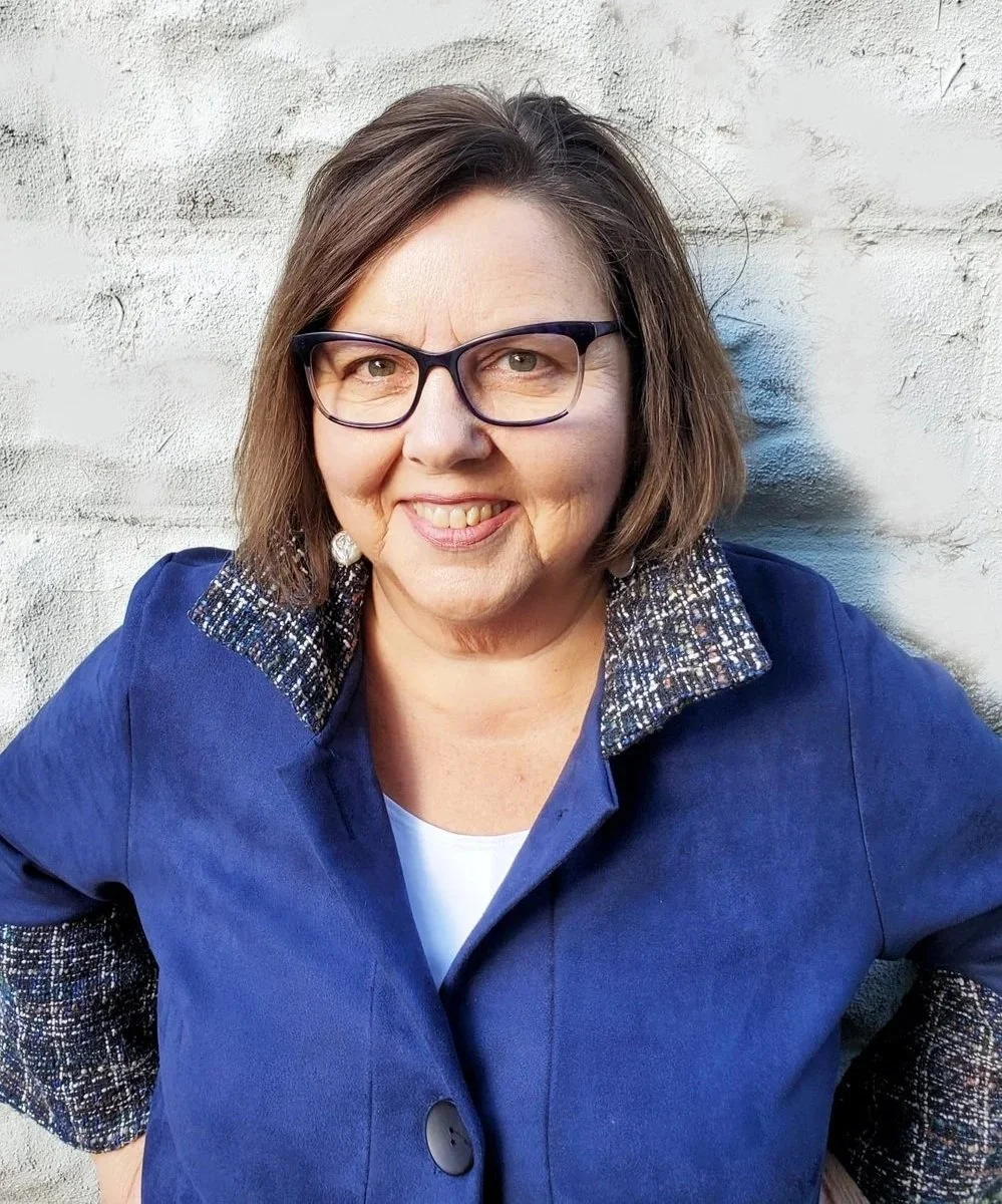 A female psychologist with short brown hair and glasses smiling, wearing a blue blazer with a patterned collar, standing outdoors against a white brick wall.