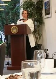 Woman speaking at a podium during a formal event with glasses and cups on a table in the foreground.