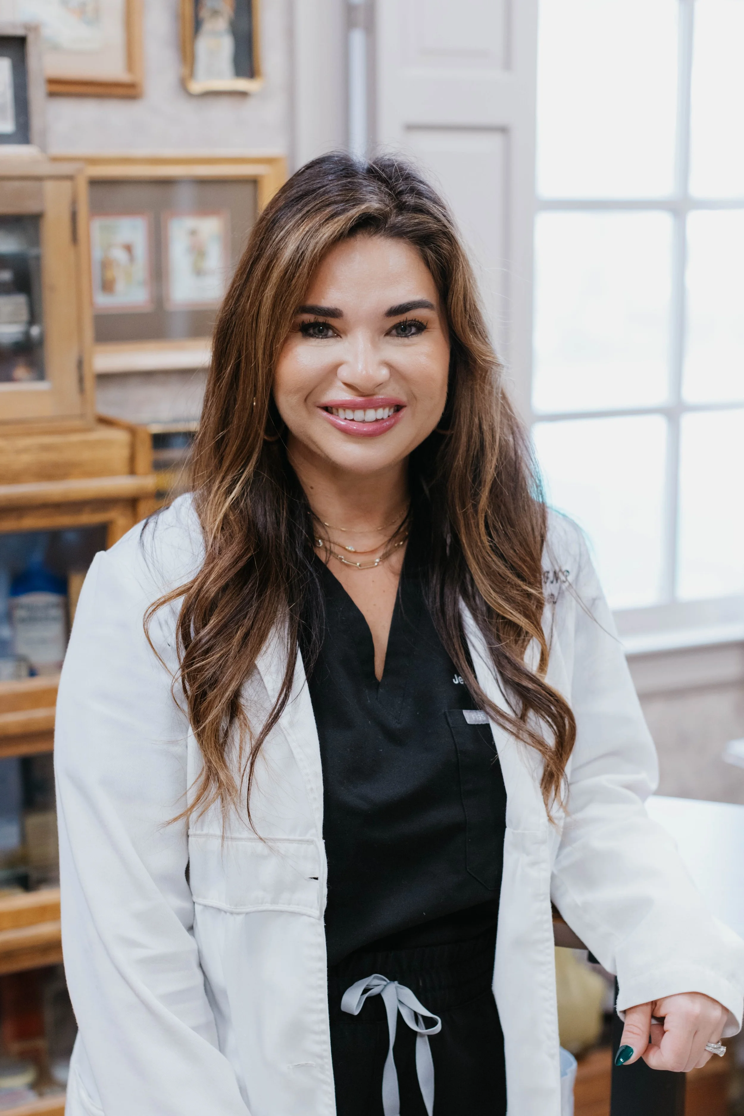 Smiling woman in a white lab coat standing in a room with framed pictures on the wall.