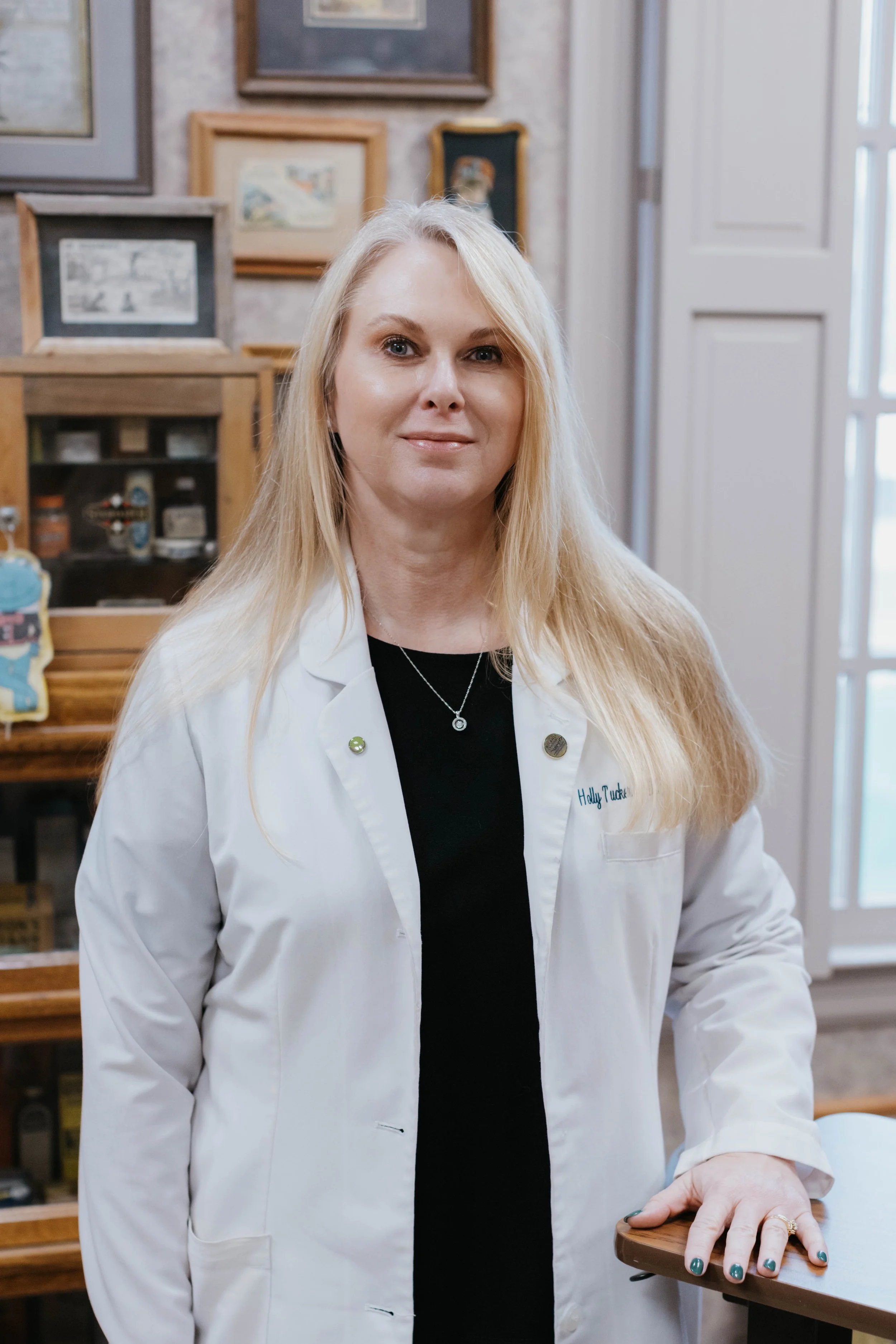 A woman wearing a white lab coat, standing in a room with framed pictures on the walls.