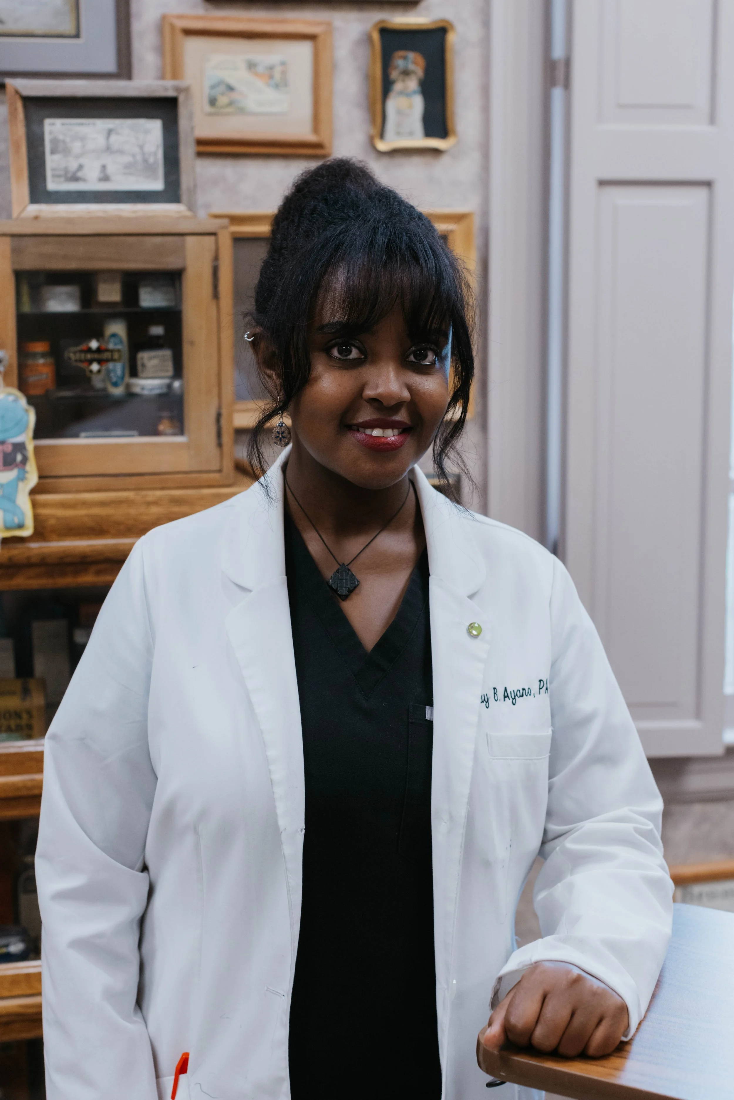 Woman in a white lab coat standing in a room with framed pictures on the wall.