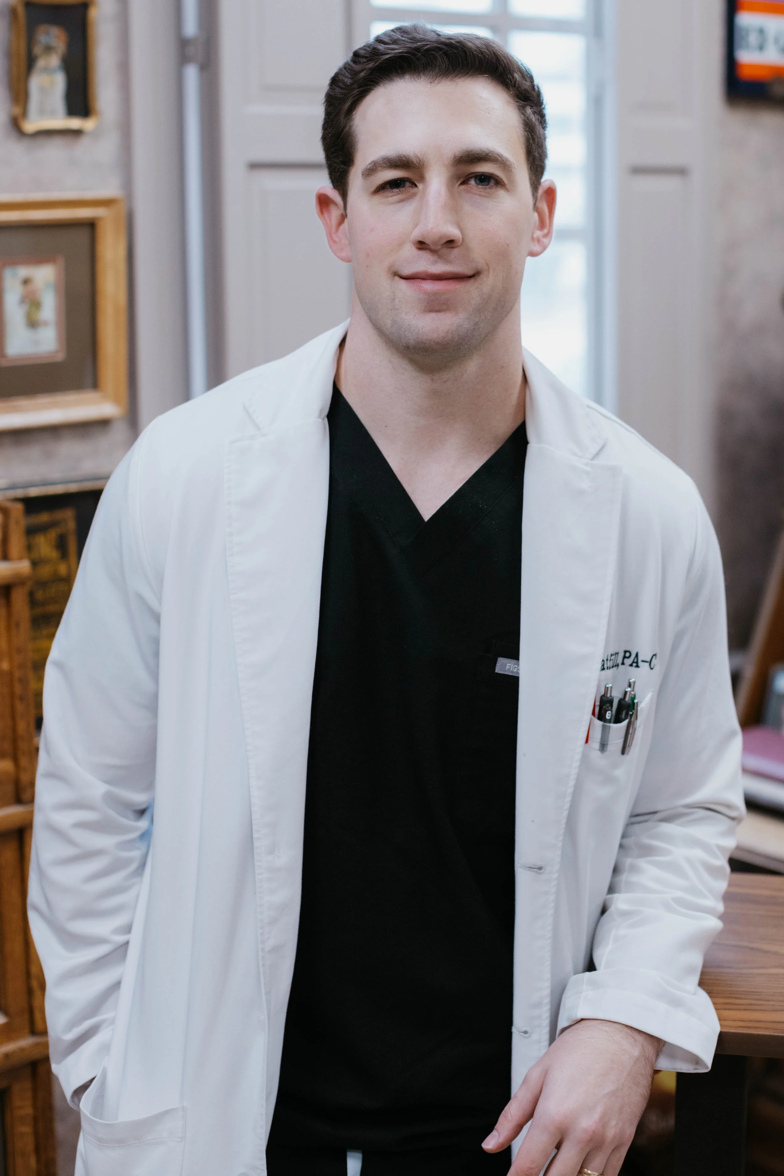 Person wearing a white lab coat and black scrubs standing indoors, with framed art on the wall in the background.