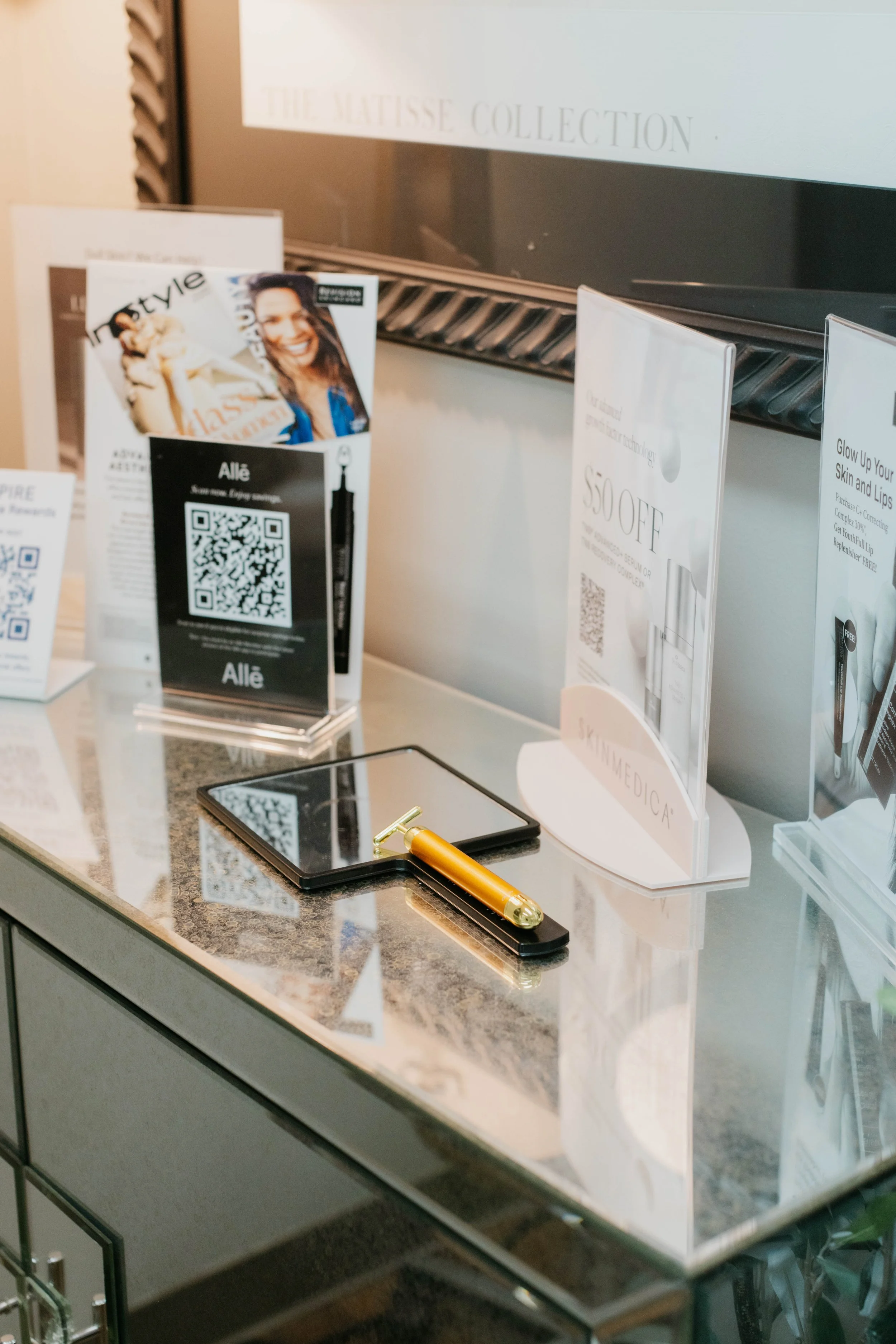 A mirror and pamphlets with QR codes on a countertop, featuring discounts and skincare products, with a background titled "The Matisse Collection."