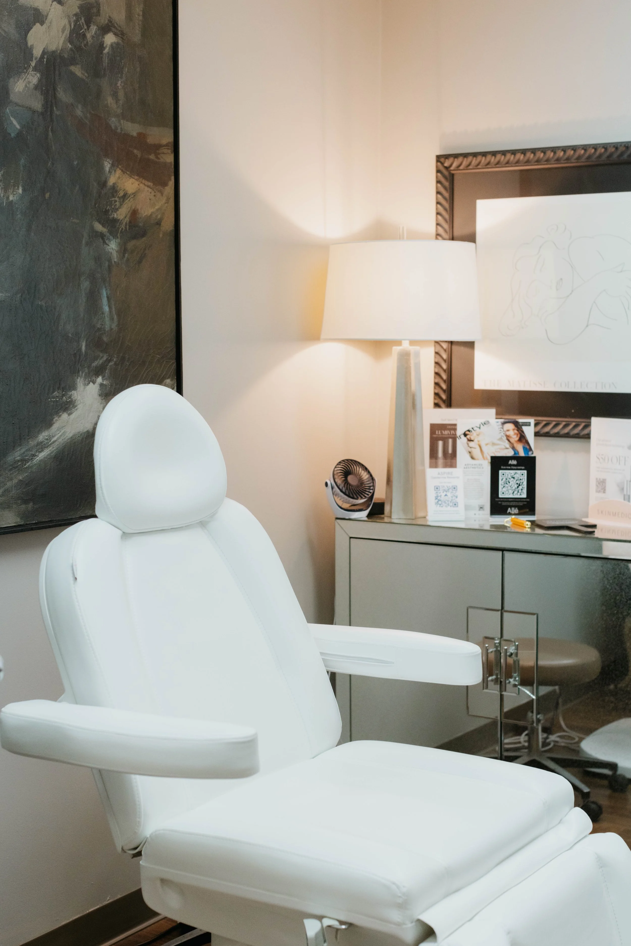 Modern spa treatment room with white reclining chair, artwork, lamp, and promotional materials on a mirrored cabinet.