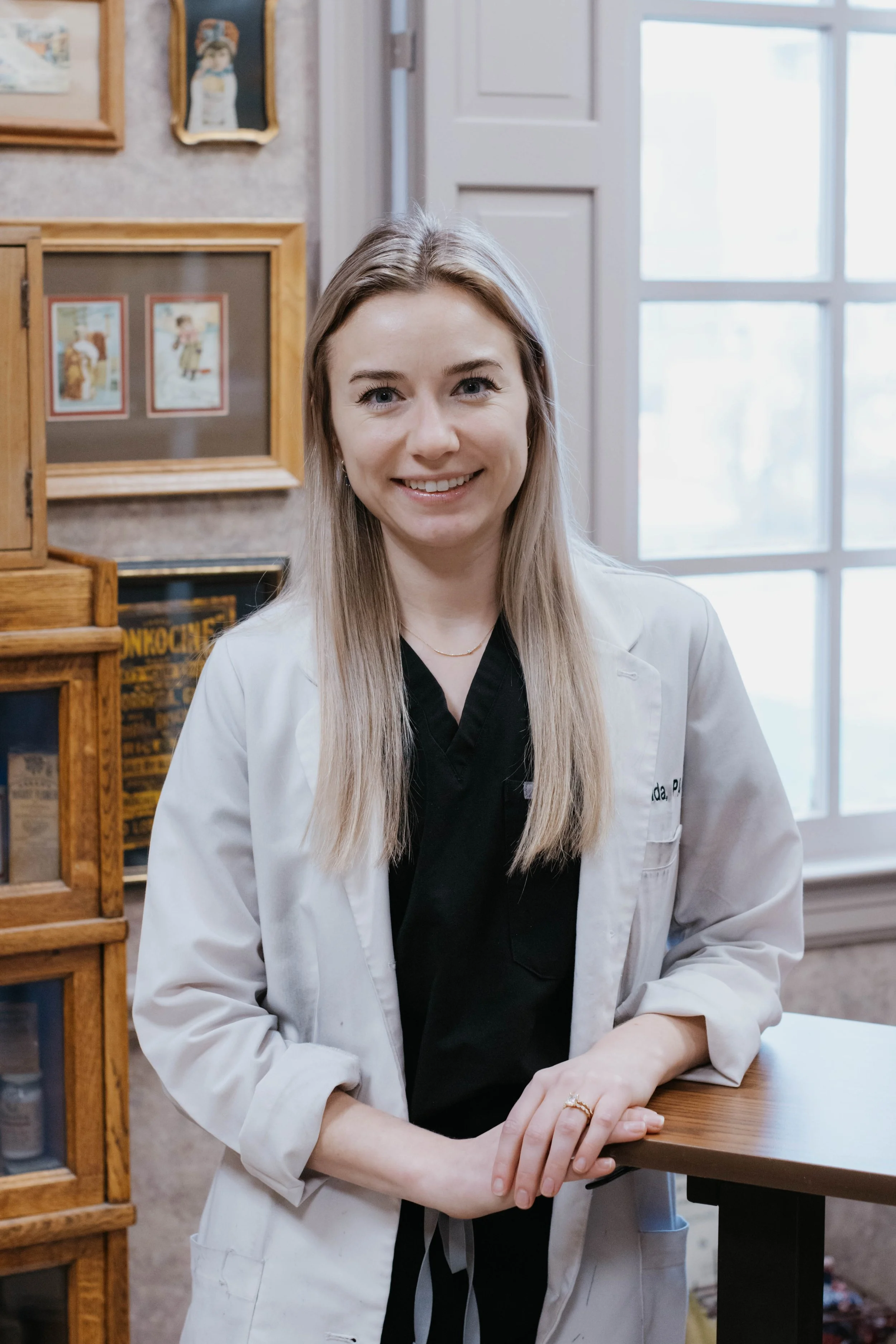 A smiling woman in a white lab coat standing indoors next to a table, with framed pictures and a window in the background.