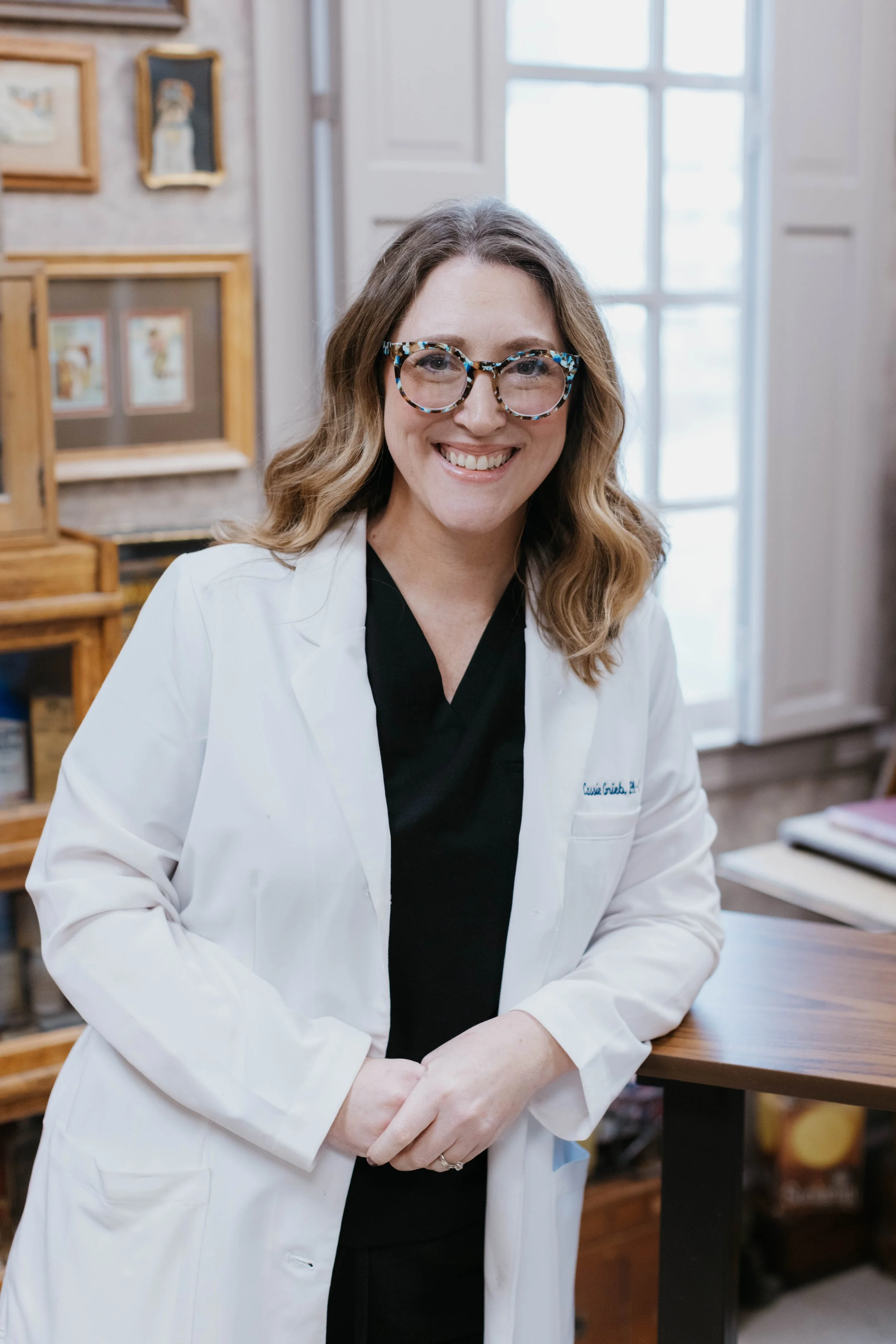 Woman in glasses wearing a white lab coat, smiling, indoors near a window with framed art on the wall.